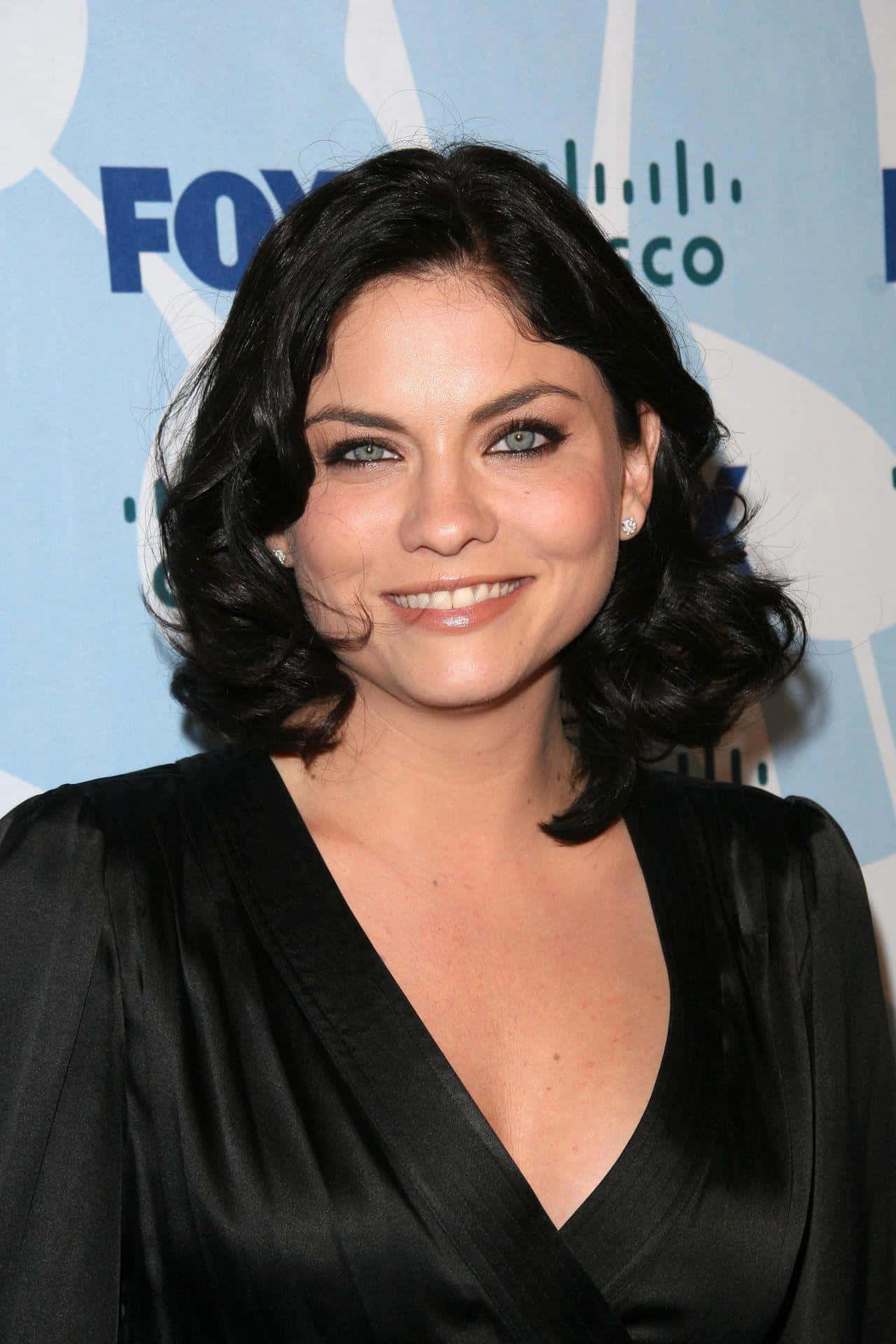 jodi lyn o'keefe 桌布 壁紙 — 一位微笑的黑髮波浪捲、綠眼睛的女性，在FOX活動中穿著黑色緞面連衣裙。