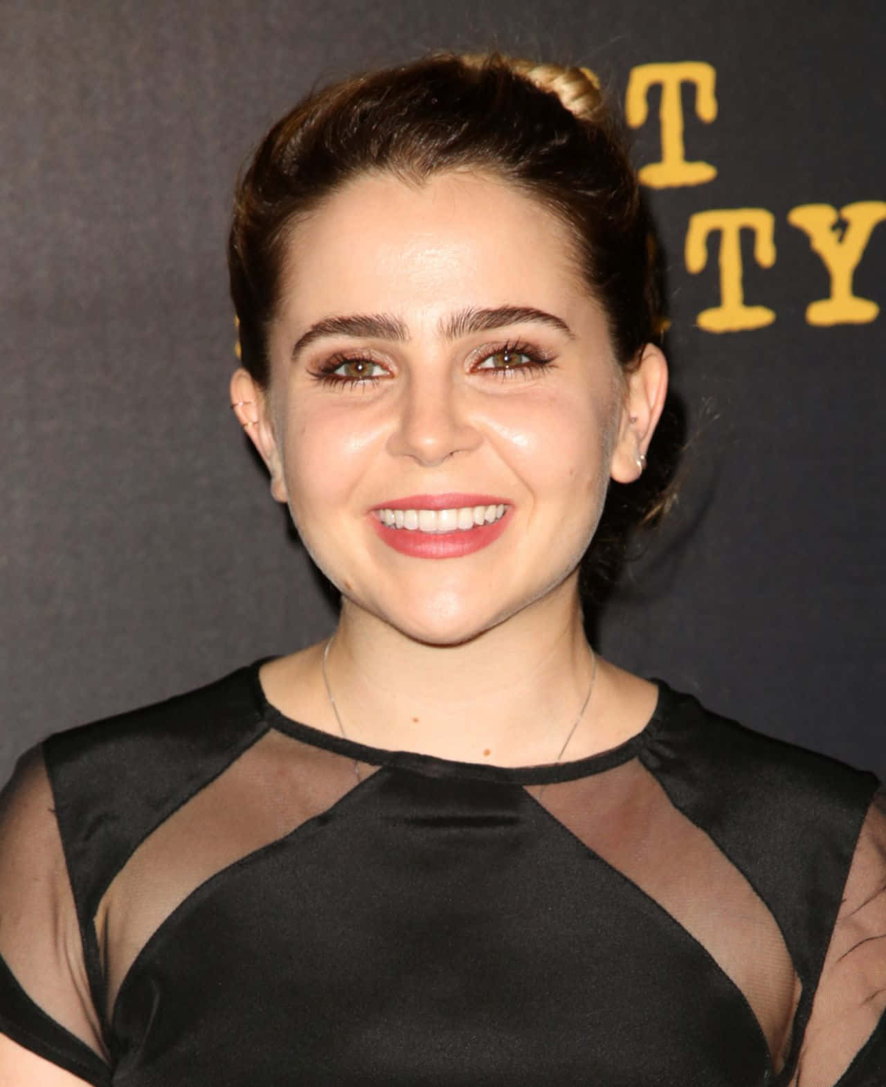 Mae Whitman 桌布 壁紙 — 一位微笑的黑髮盤起女子，身穿黑色透明袖連衣裙，背景為深色並帶有黃色字樣。