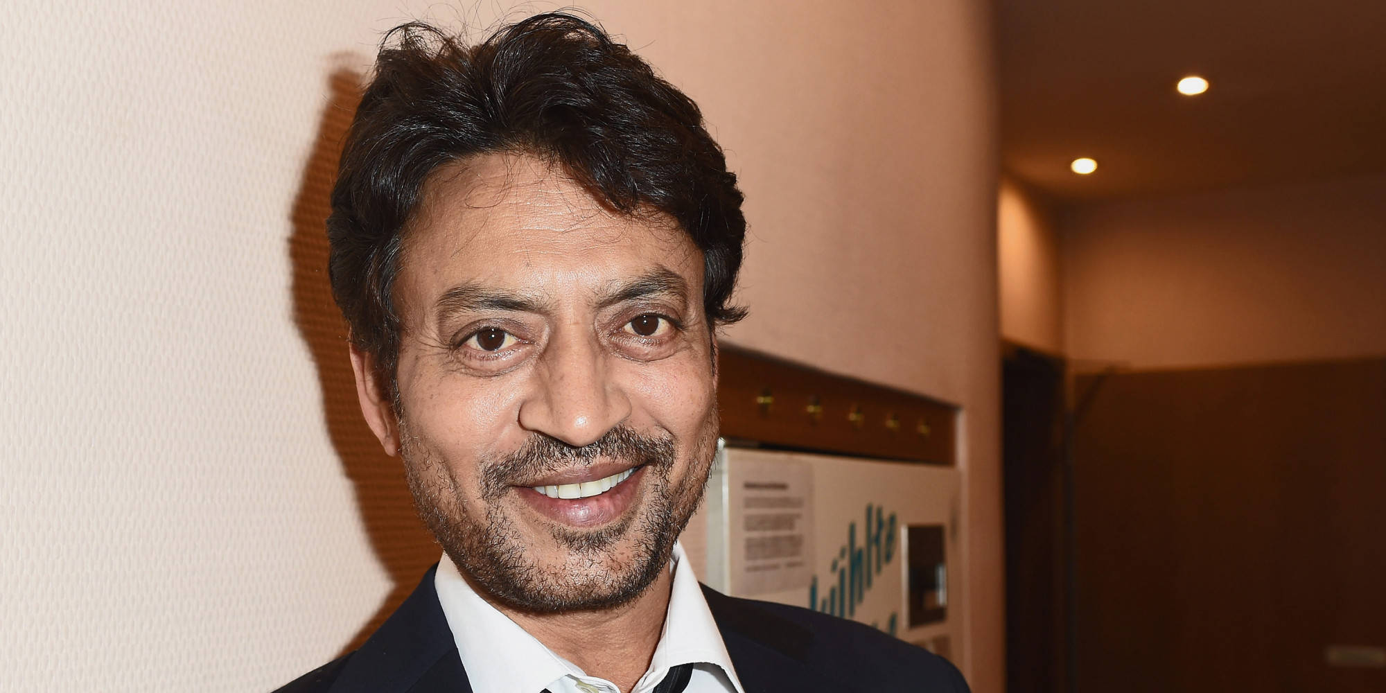 Irrfan Khan 桌布 壁紙 — 一位微笑的黑髮鬍鬚男士，在室內暖光下穿著深色西裝和白色襯衫。