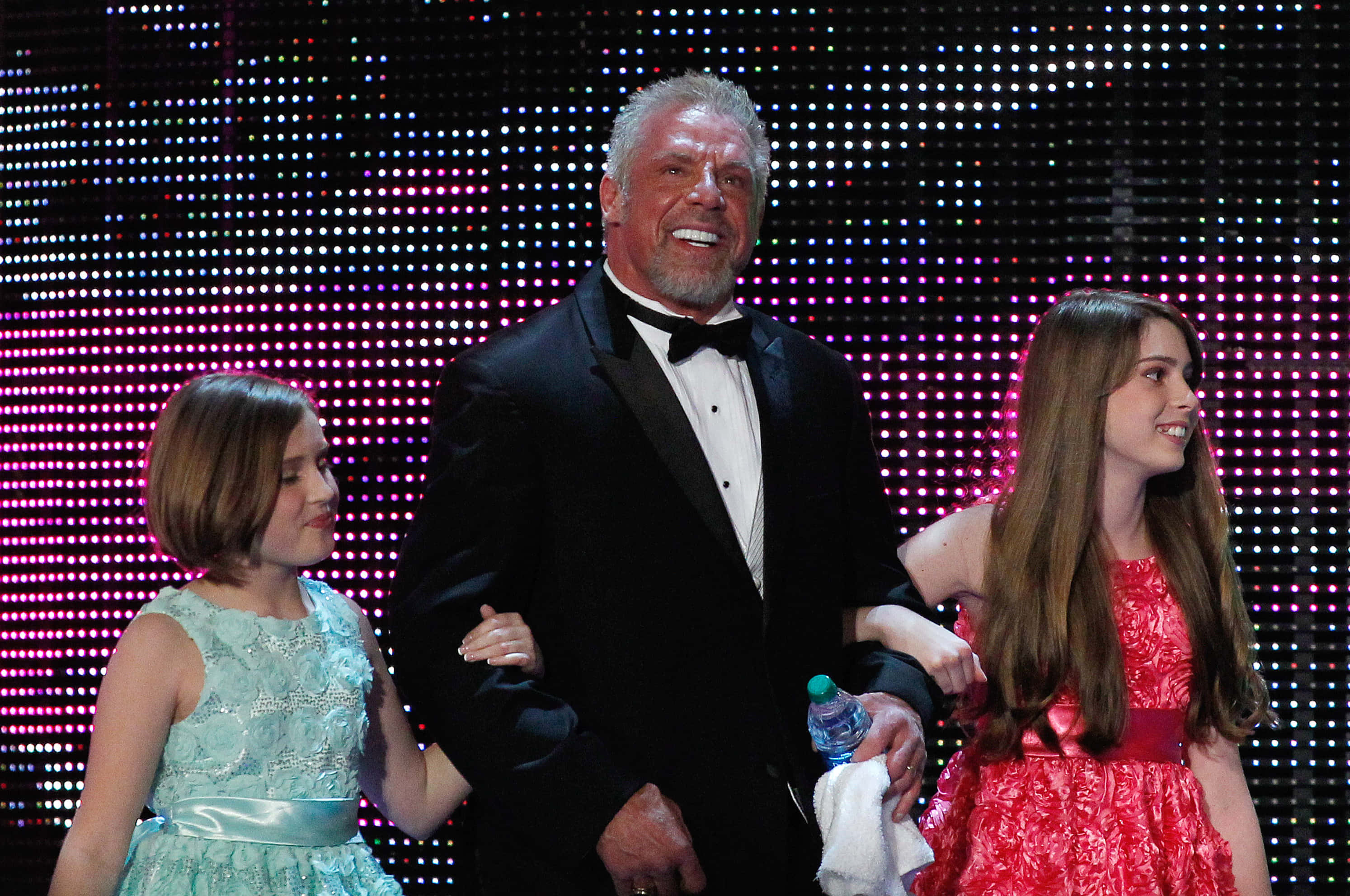 WWE 桌布 壁紙 — 一位微笑身穿燕尾服的男士站在兩個年輕女孩之間，舞台燈光明亮，背景是像素化LED螢幕。