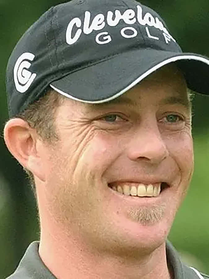 高爾夫 桌布 壁紙 — 一位微笑高爾夫球手的特寫肖像，戴著黑色 Cleveland Golf 帽子，背景是模糊的綠色。