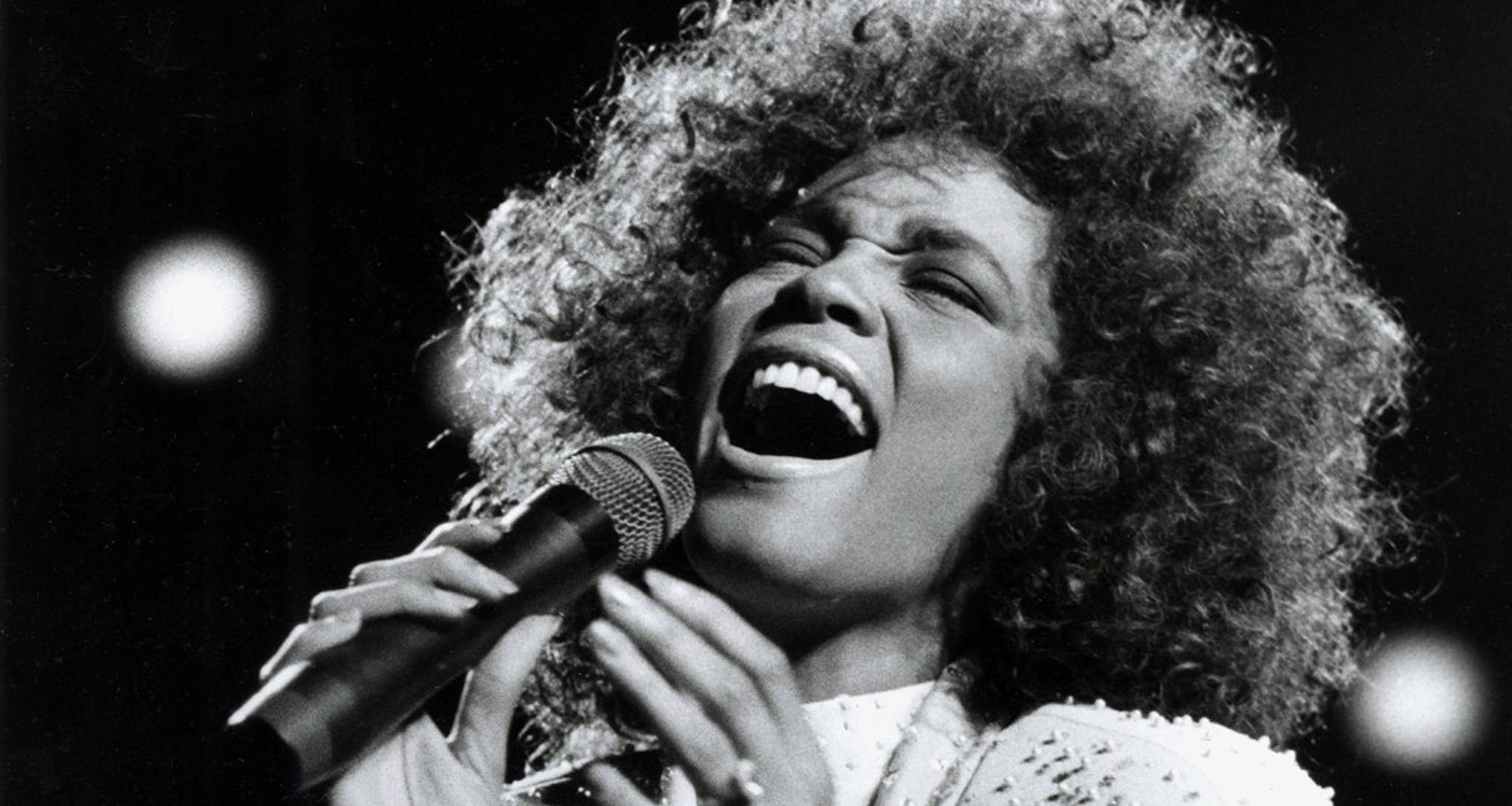 Whitney 桌布 壁紙 — 一位快樂歌手的黑白特寫，他有豐盈捲髮，在舞台燈光下熱情地對著麥克風唱歌。