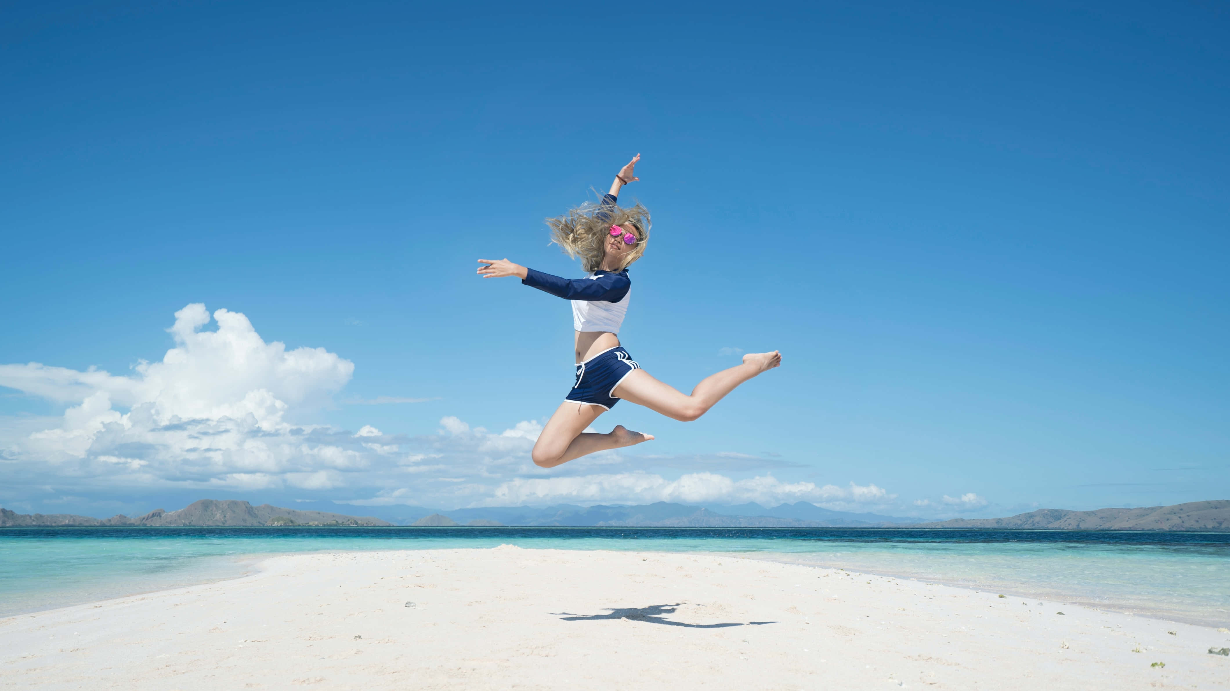 Summer Vacation 桌布 壁紙 — 一位快樂的女人在廣闊藍天和蓬鬆白雲下，於純淨白沙灘上高高躍起。