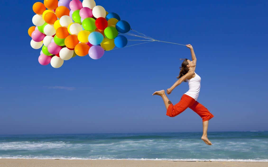 Balloon 桌布 壁紙 — 一位快樂的女人在陽光明媚的海灘上高高跳起，一簇色彩繽紛的氣球將她托起，映襯著清澈的藍天。