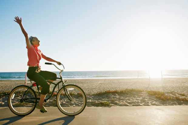 bike riding 桌布 壁紙 — 一位快樂的女人在陽光明媚的海灘上騎著腳踏車，一隻手臂舉向明亮的地平線。