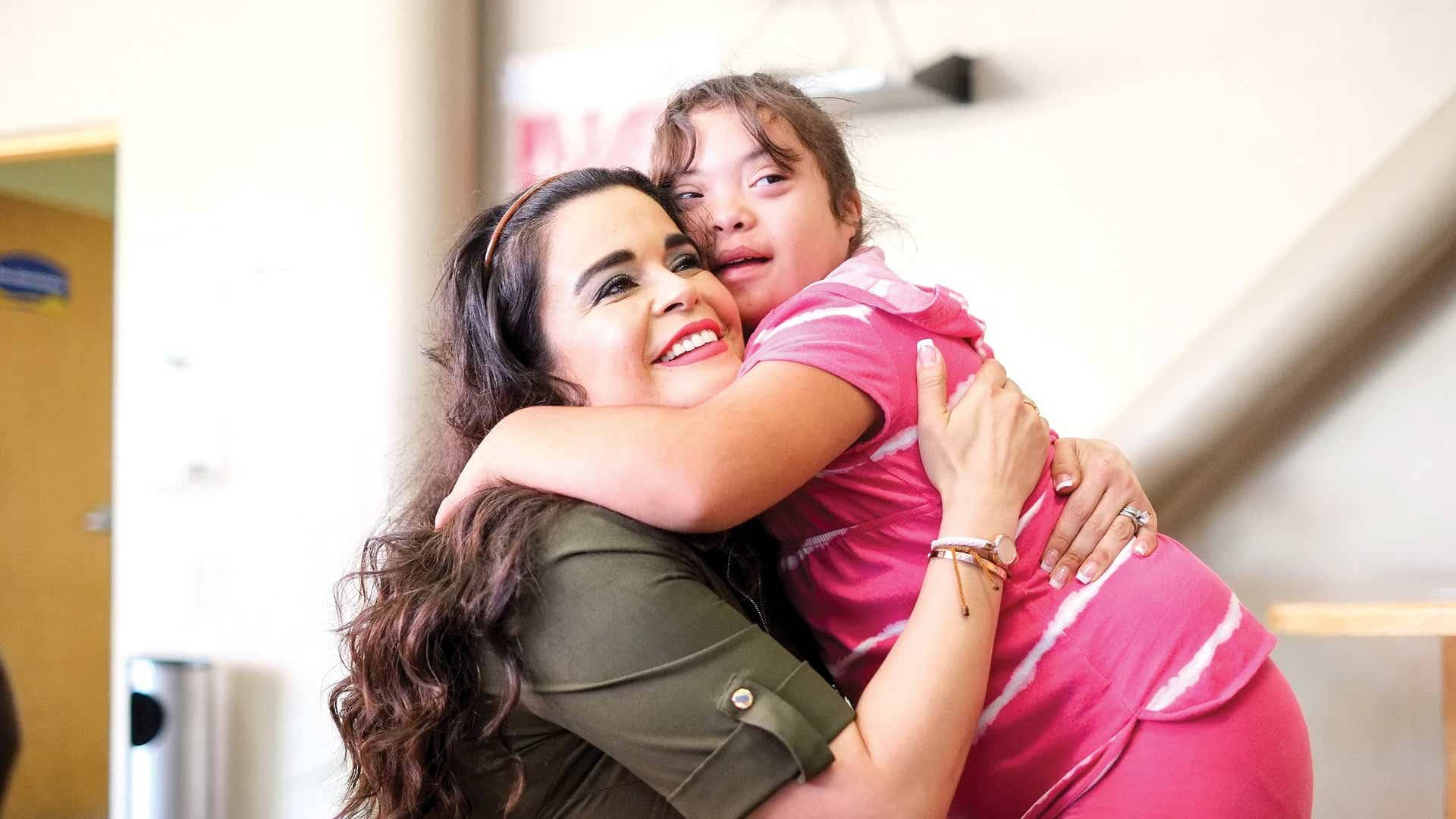 Hug 桌布 壁紙 — 一位快樂的女人在陽光照射的室內走廊裡，帶著溫暖的笑容擁抱著一個穿著亮粉紅色襯衫的小女孩。