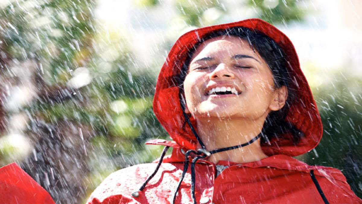下雨 桌布 壁紙 — 一位快樂的女人穿著紅色雨衣，在陽光明媚的戶外場景中，閉著眼睛大笑，雨水如瀑布般灑落在她周圍。