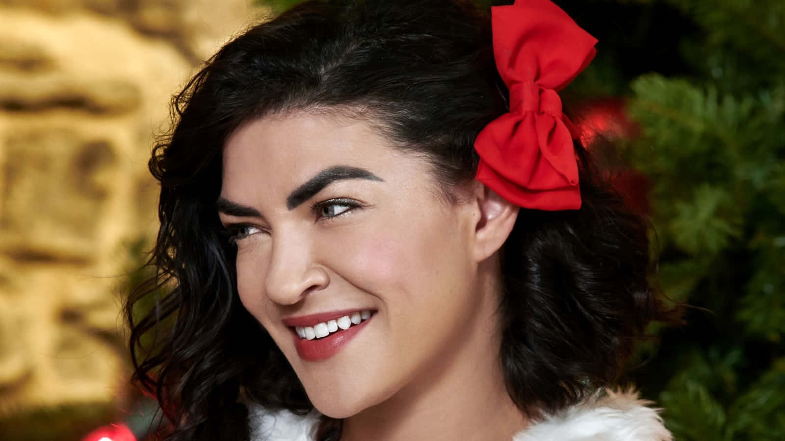 jessica szohr 桌布 壁紙 — 一位快樂的女士，深色捲髮上戴著紅色蝴蝶結，在節日綠色和金色背景前溫暖微笑。