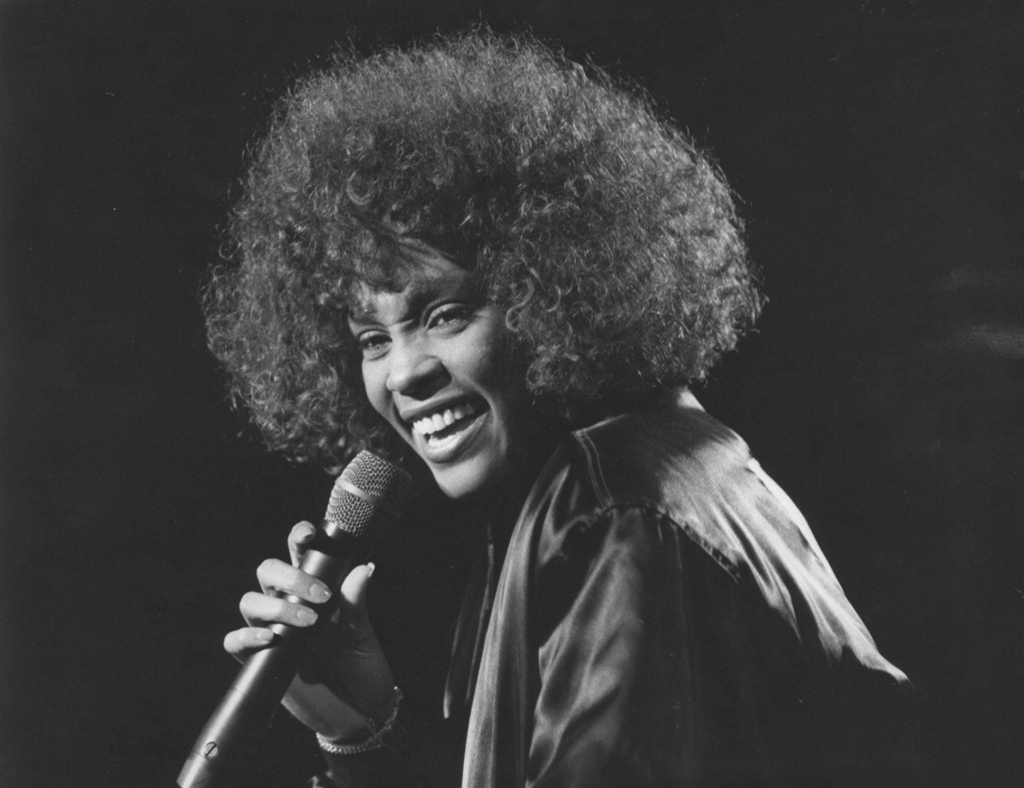 Whitney 桌布 壁紙 — 一位快樂的女士，留著濃密的非洲式爆炸頭，在黑暗背景下拿著麥克風大笑。
