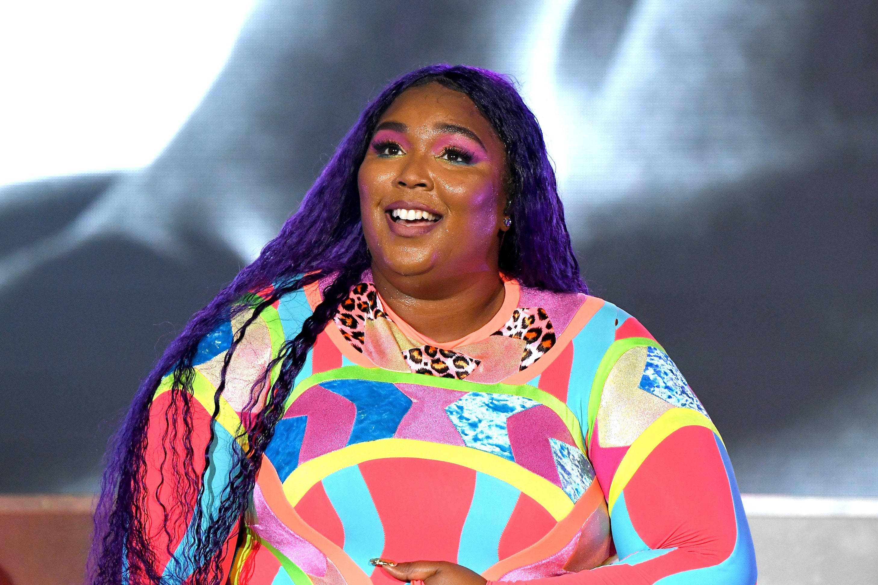 Lizzo 桌布 壁紙 — 一位快樂的女士，紫色頭髮，穿著鮮豔多彩的服裝，在戲劇性燈光下於舞台上燦爛微笑。