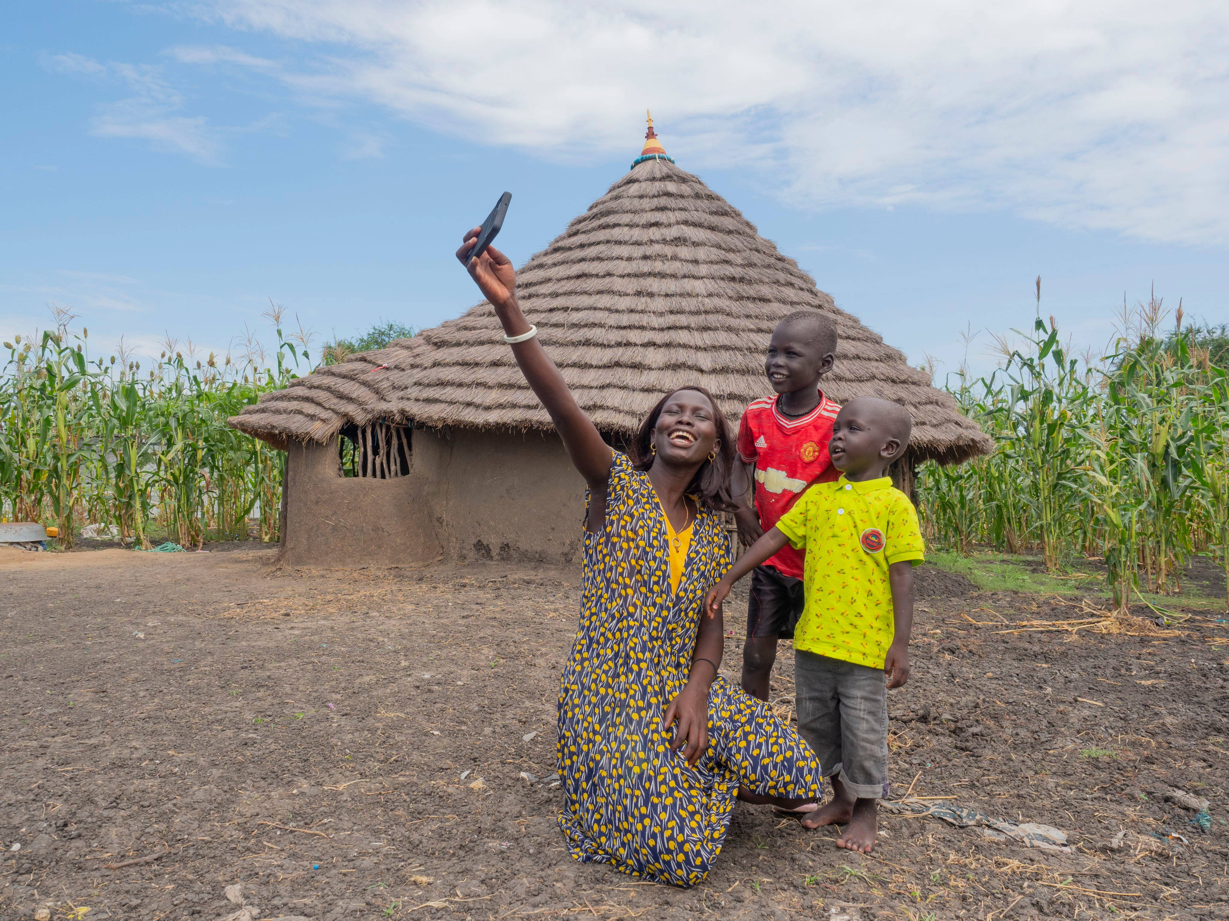 South Sudan 桌布 壁紙 — 一位快樂的女士和兩個孩子在藍天白雲下，於高聳的玉米田環繞的茅草屋外自拍。