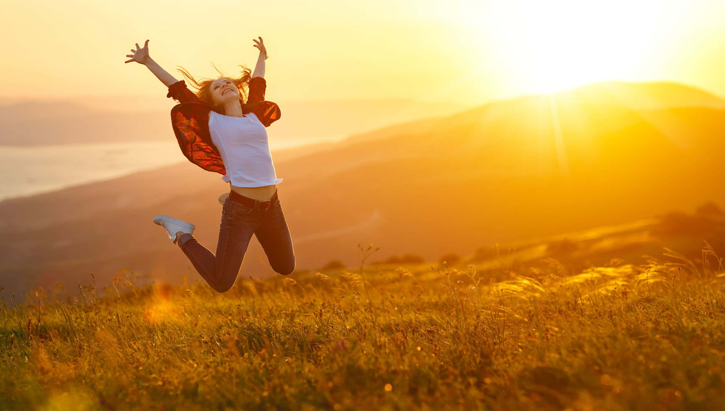 Contentment 桌布 壁紙 — 一位快樂的女子在金色田野中跳躍，夕陽在起伏的山丘後落下，灑下溫暖的光芒。