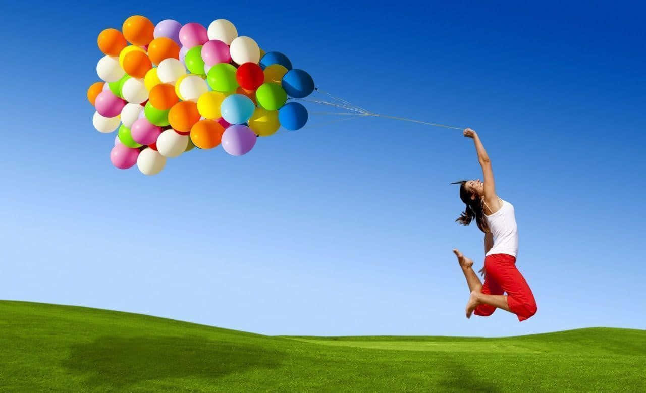 happiness 桌布 壁紙 — 一位快樂的女性在一片充滿活力的綠色田野中跳躍，一簇色彩繽紛的氣球將她托向藍天，背景清澈蔚藍。