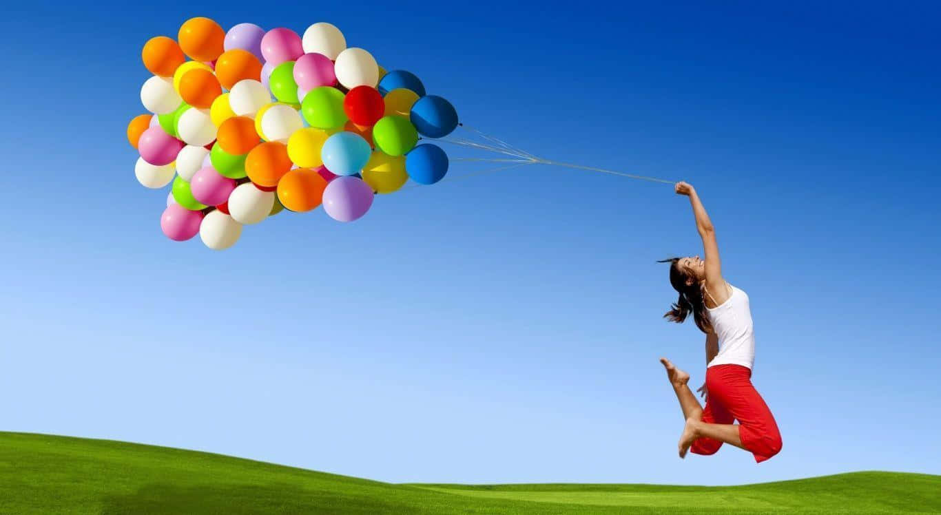 happiness 桌布 壁紙 — 一位快樂的女性在晴朗的藍天下向上跳躍，被一束色彩繽紛的氣球拉著，下方是翠綠的田野。