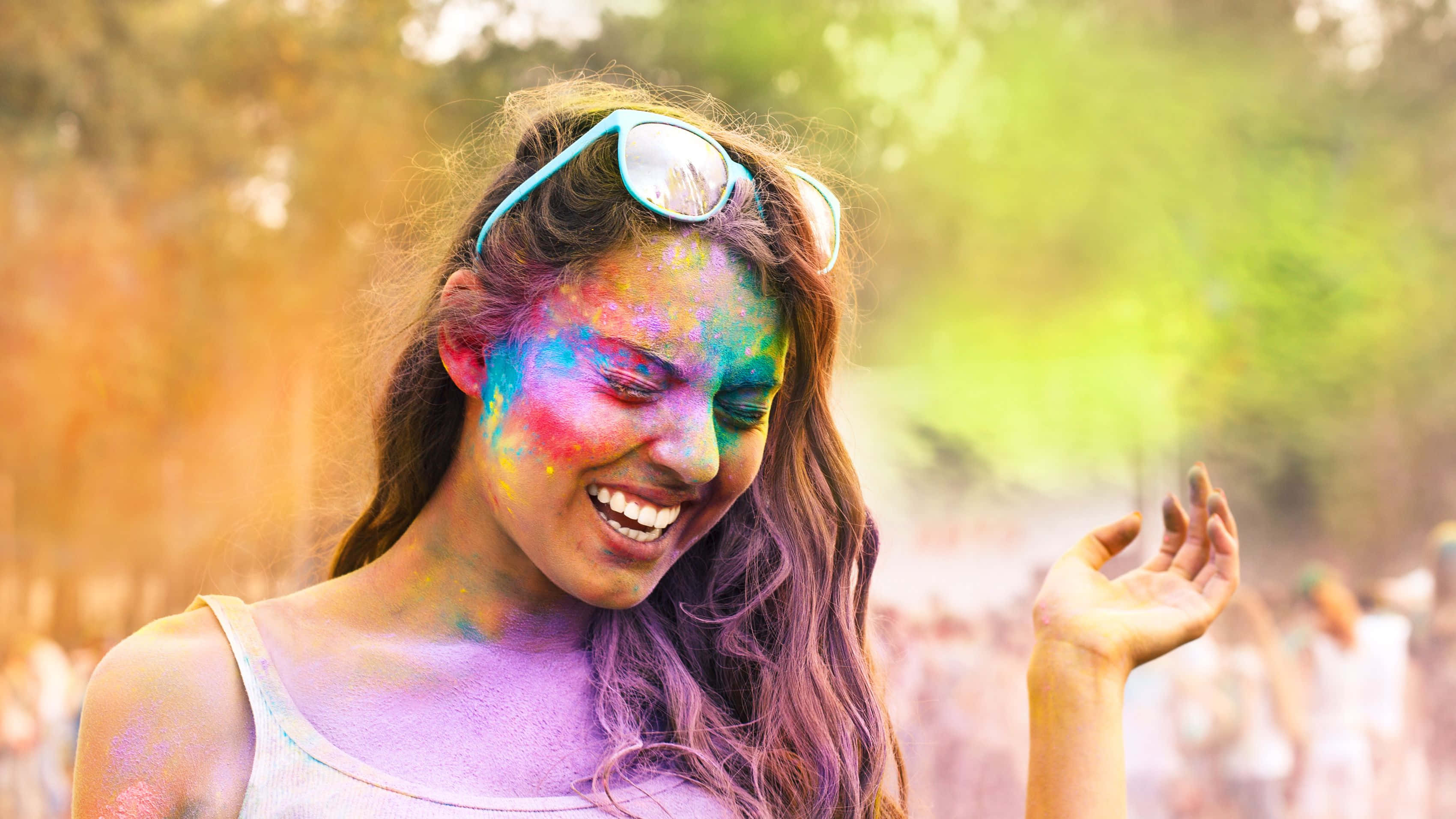 Holi 桌布 壁紙 — 一位快樂的女性在模糊的戶外背景下，臉上和頭髮上沾滿鮮豔的彩色粉末，開懷大笑。