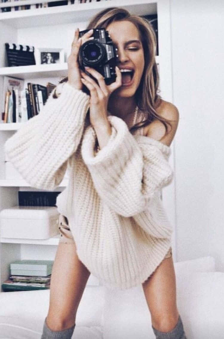Holding Camera 桌布 壁紙 — 一位快樂的女性穿著舒適的露肩奶油色毛衣，在明亮的白色房間裡笑著，同時將一部復古相機舉到臉前。