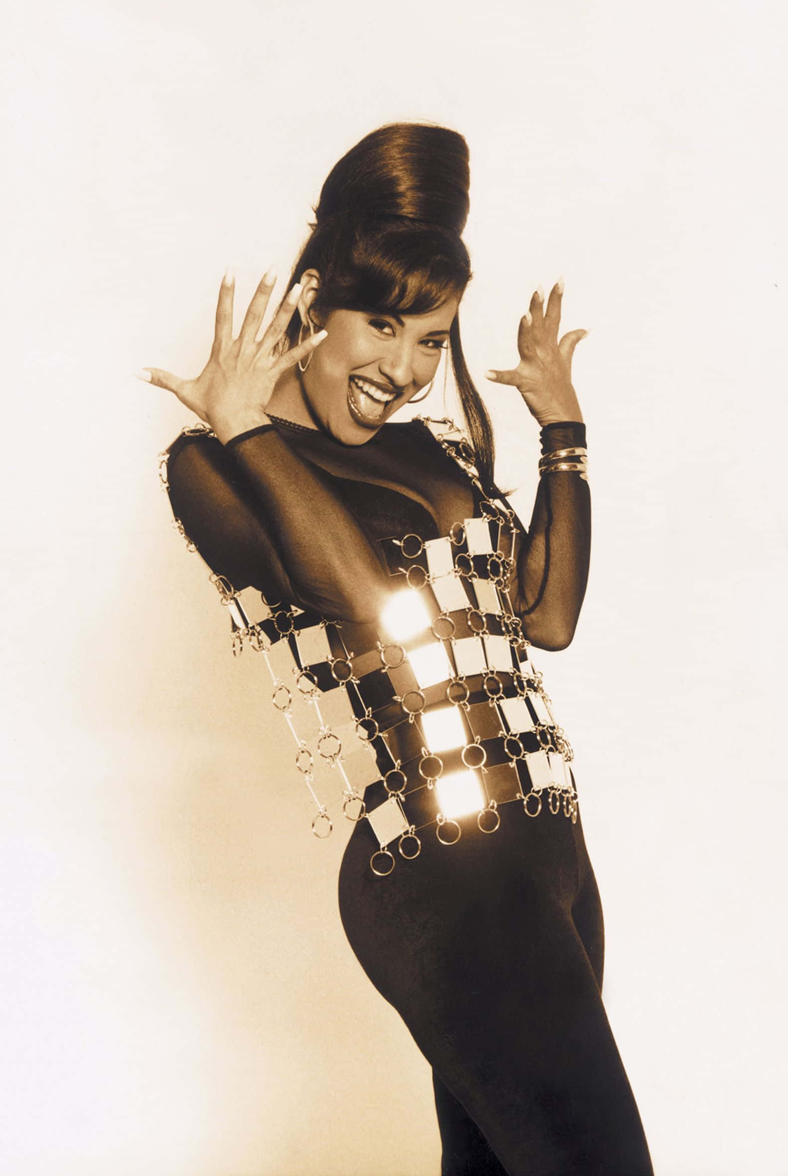 Selena Quintanilla iPhone 桌布 壁紙 — 一位快樂的女性穿著閃亮的金屬鏈服裝，在純色背景前舉起雙手擺姿勢。