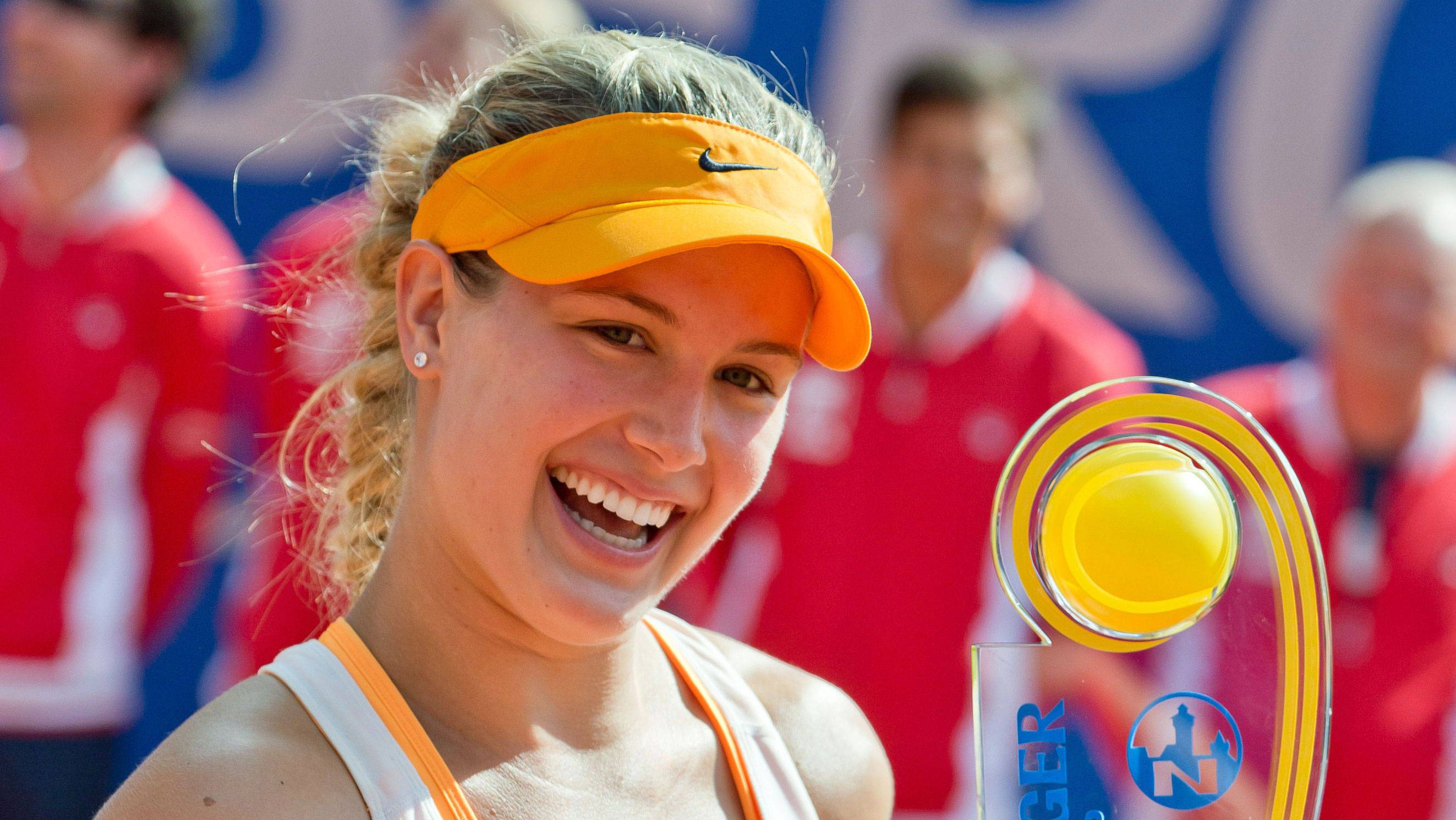 Eugenie Bouchard 桌布 壁紙 — 一位快樂的女網球運動員在明亮的陽光下，手持一個黃色透明的獎盃，笑容燦爛。