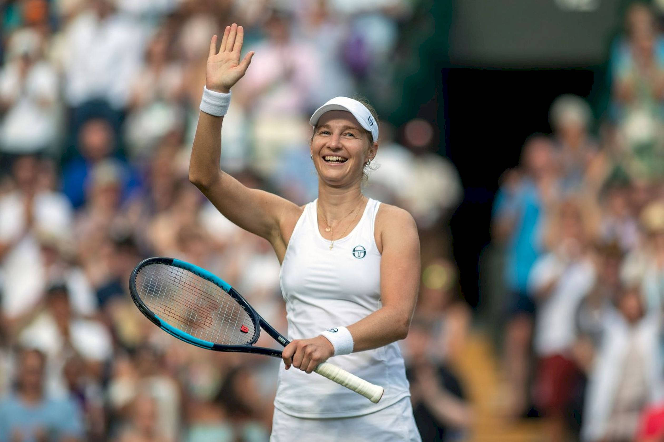 運動 桌布 壁紙 — 一位快樂的女網球選手在陽光照耀的草地場上，一邊握著球拍一邊向歡呼的觀眾揮手。