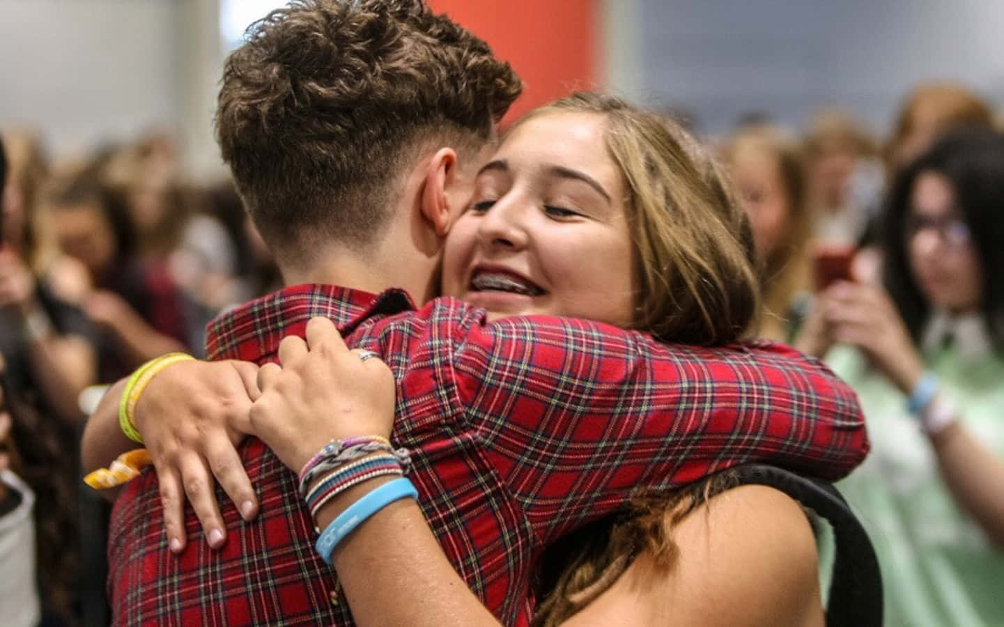 Hug 桌布 壁紙 — 一位快樂的年輕女子在擁擠的活動中閉著眼睛笑著擁抱一個穿著紅色格紋襯衫的男孩。