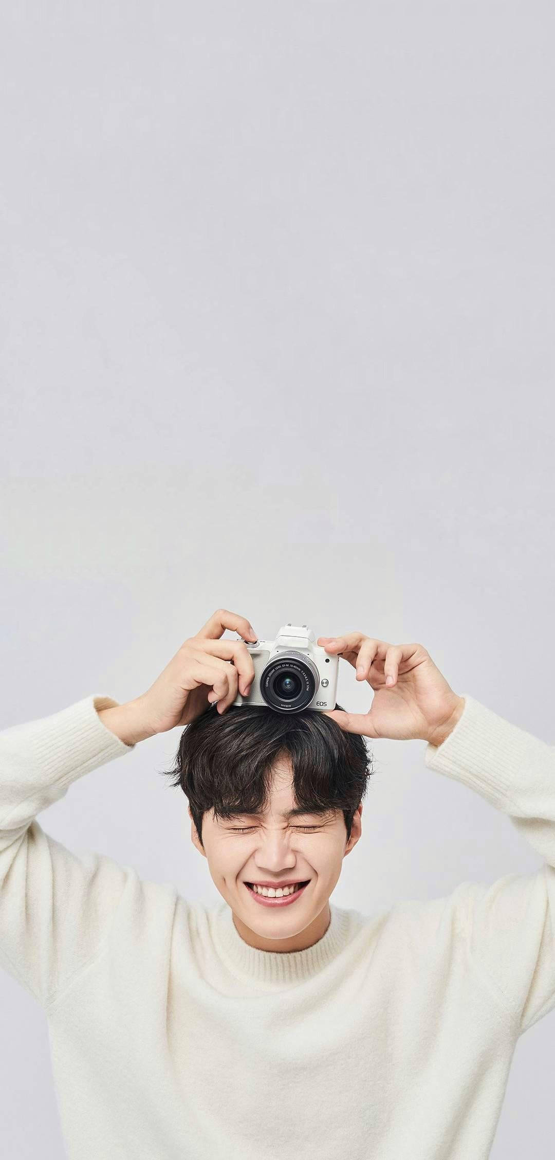 Kim Seon Ho 桌布 壁紙 — 一位快樂的年輕男子穿著奶油色毛衣，在淺灰色背景上頑皮地將白色相機平衡在頭上。