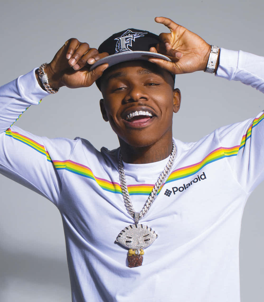 Dababy 桌布 壁紙 — 一位快樂的年輕男子穿著白色Polaroid襯衫，帶有彩虹條紋和閃閃發光的杯子蛋糕吊墜，在純灰色背景前微笑著調整帽子。