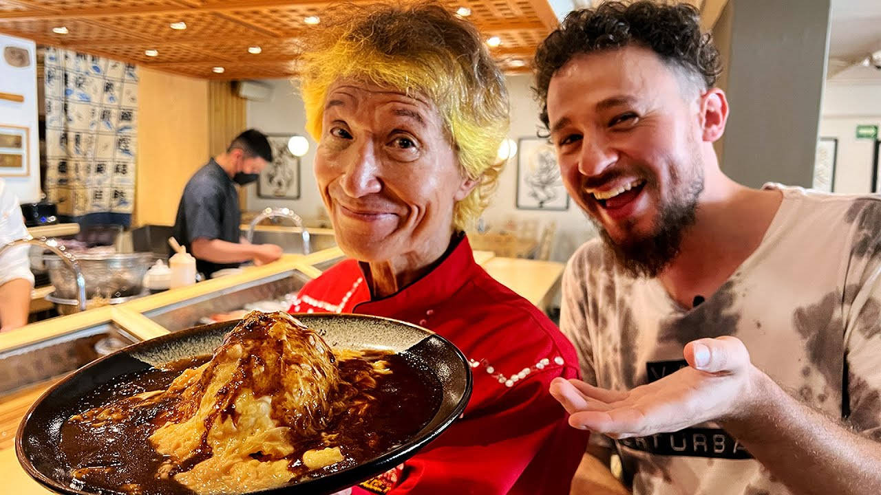 烹飪 桌布 壁紙 — 一位快樂的廚師和客人在溫暖的餐廳環境中，自豪地微笑著站在一座高聳的巧克力覆蓋的甜點旁。