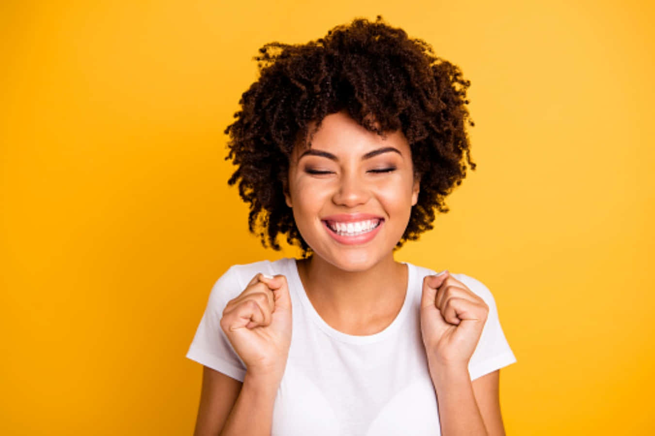 Happy Face 桌布 壁紙 — 一位快樂的捲髮女性，閉著眼睛、握緊拳頭，在鮮黃色背景前微笑。