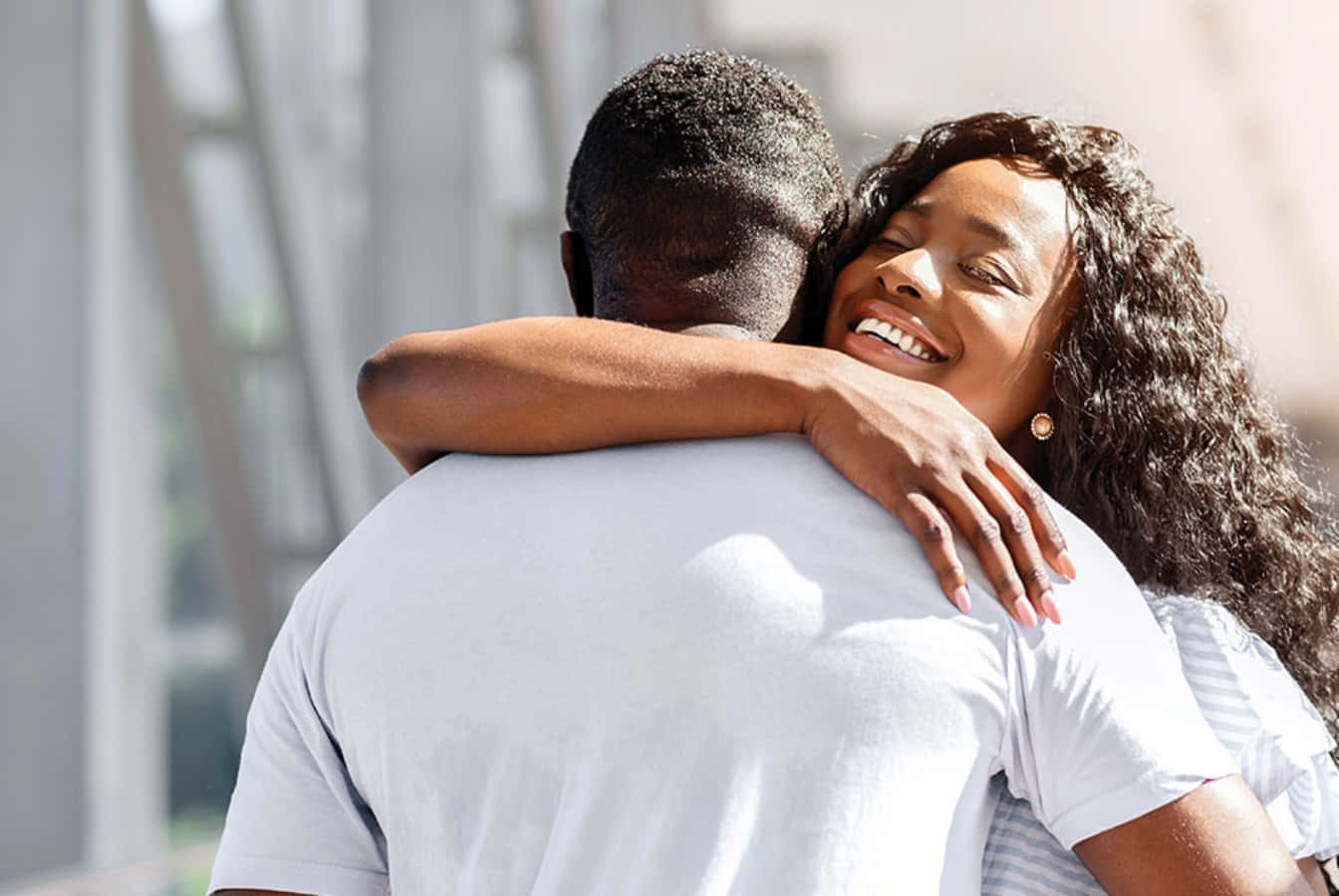 Hug 桌布 壁紙 — 一位快樂的捲髮女性在現代建築附近明亮的陽光下從後面擁抱一名男子。