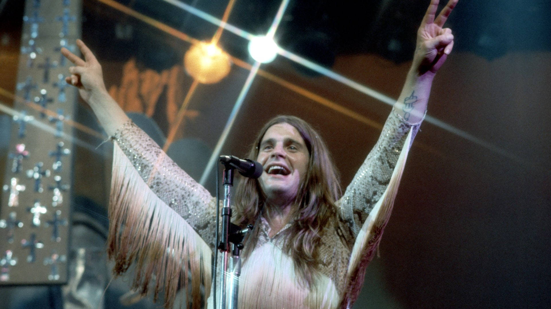 Ozzy Osbourne 桌布 壁紙 — 一位快樂的搖滾歌手，長髮飄逸，穿著流蘇閃亮袖子，在明亮的聚光燈下在舞台上表演，雙手舉起和平手勢。