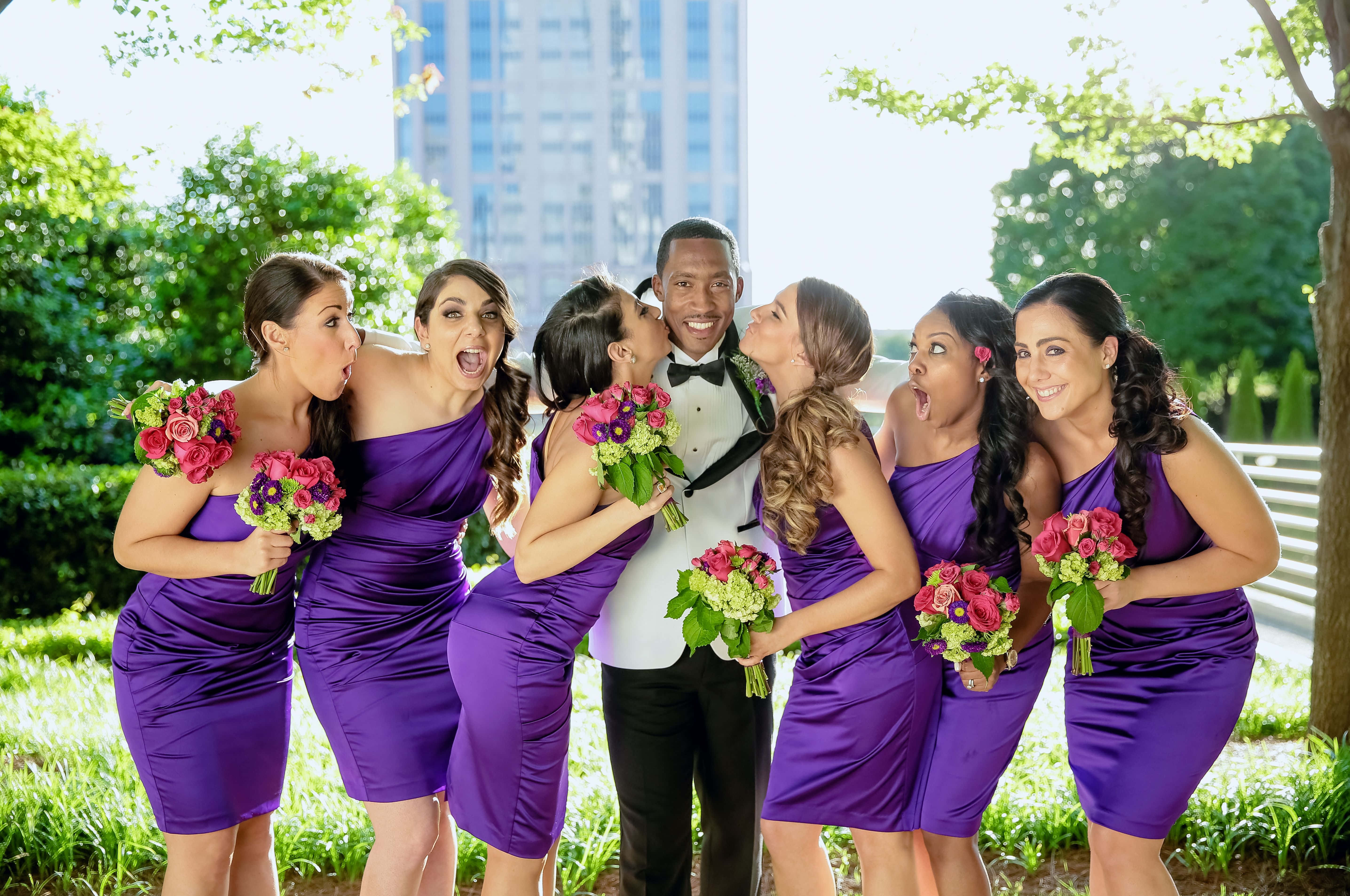 Bridesmaids 桌布 壁紙 — 一位快樂的新郎被穿著紫色連衣裙的伴娘包圍，在戶外頑皮地親吻他的臉頰。
