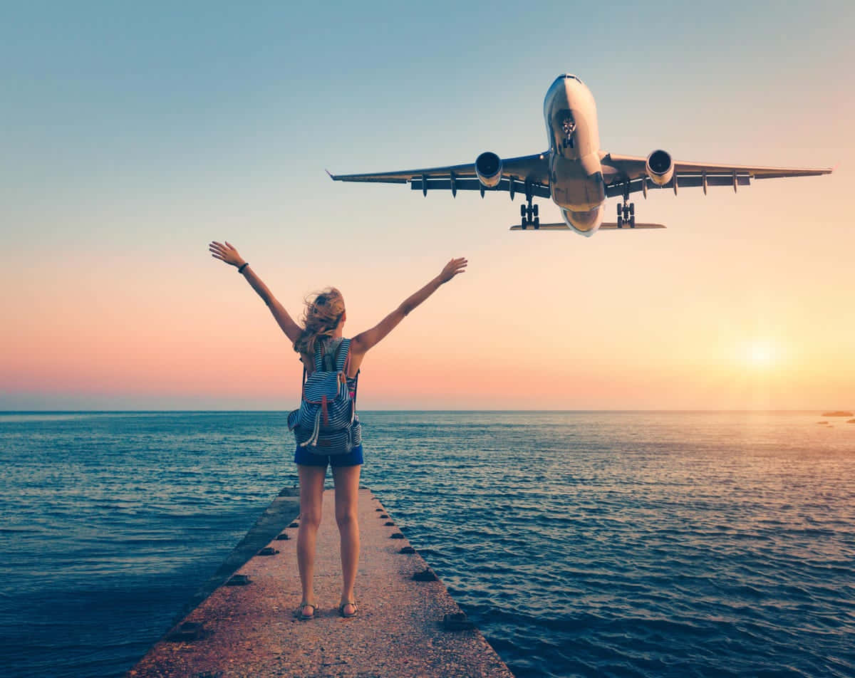 Travel Desktop 桌布 壁紙 — 一位快樂的旅行者高舉雙臂站在碼頭上，夕陽西下，一架大型飛機正在海洋上空下降準備著陸。
