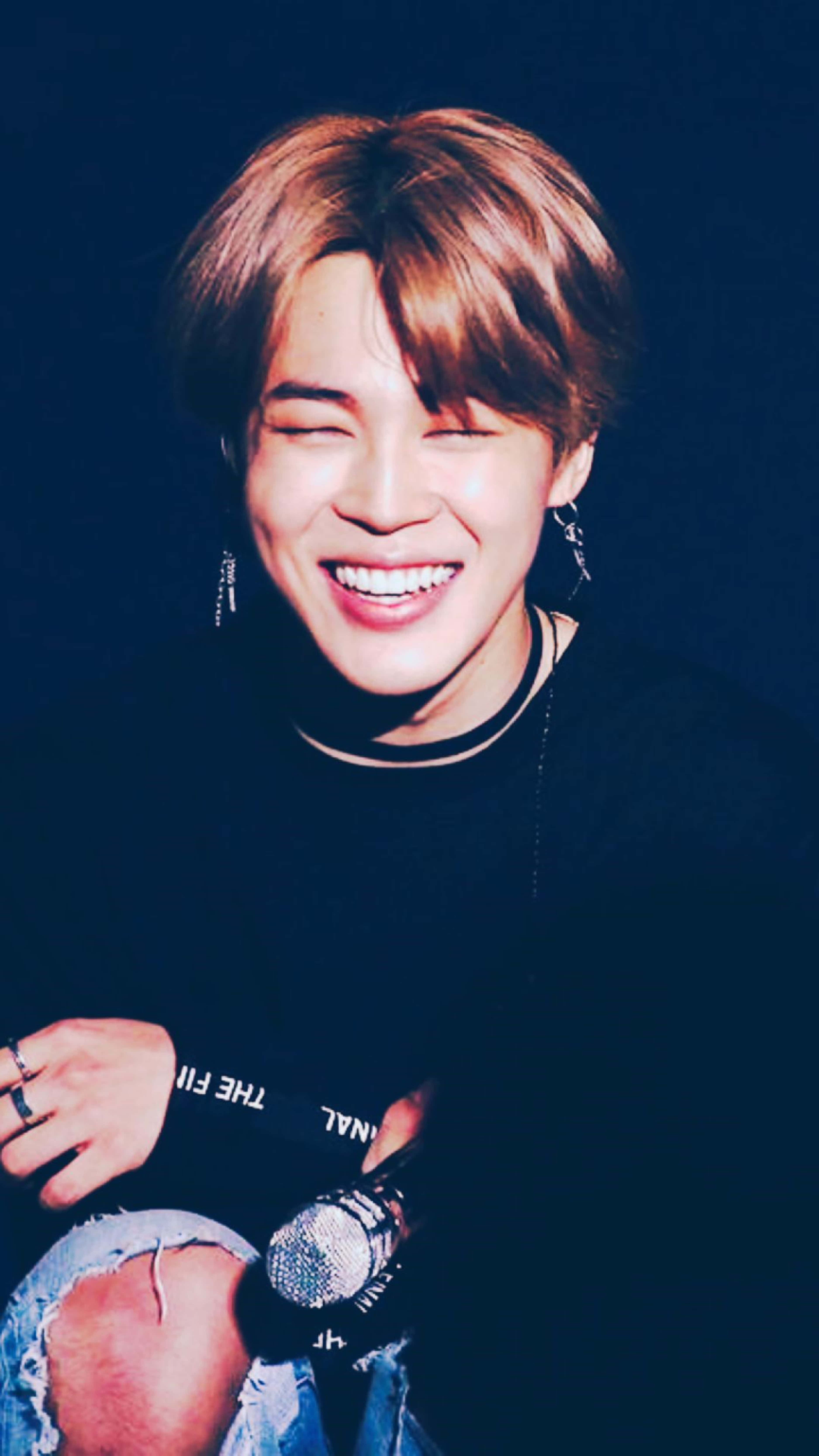 BTS 智旻 桌布 壁紙 — 一位快樂的棕髮年輕男子在黑暗背景下拿著麥克風眨眼。