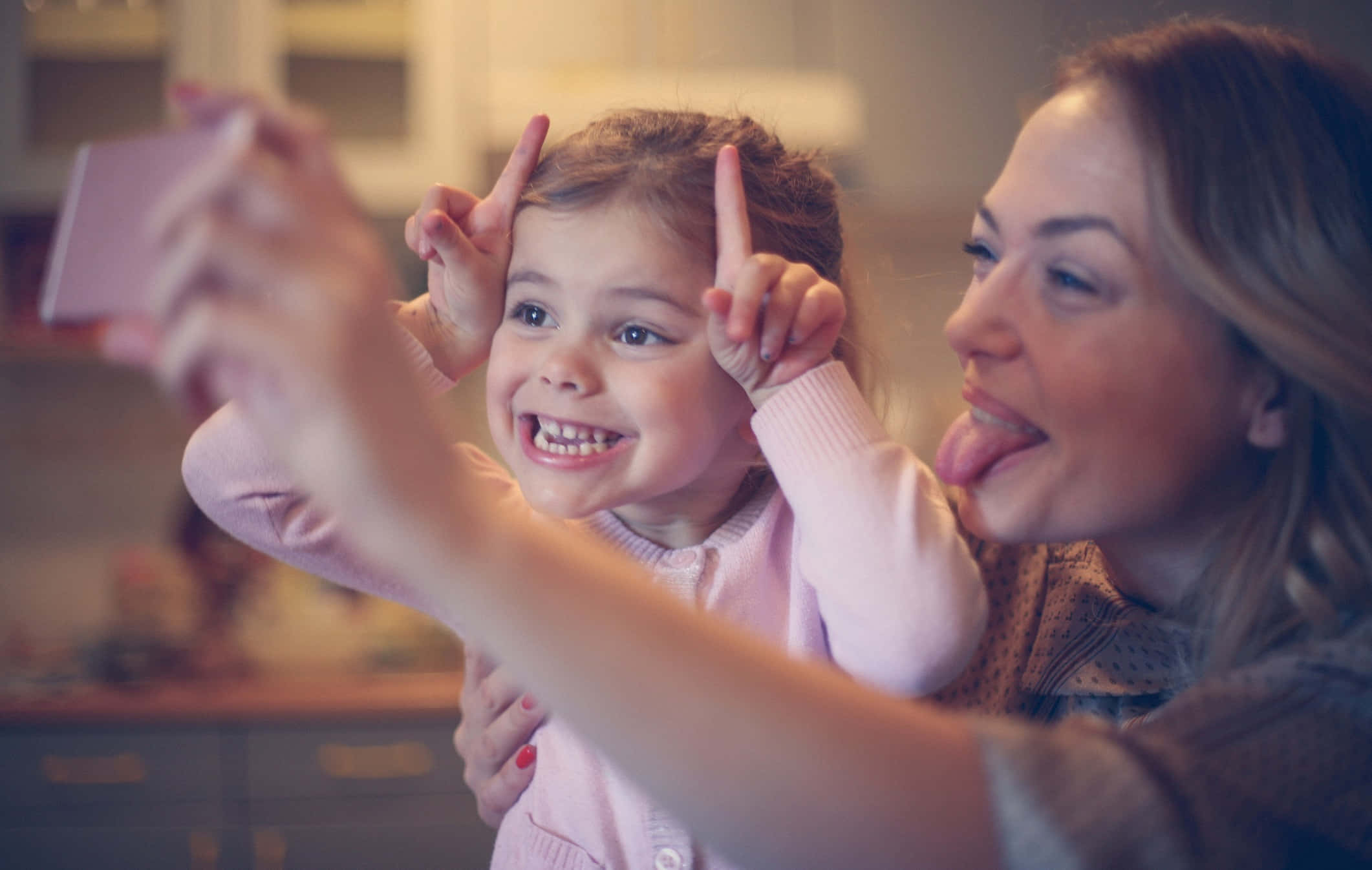 Family 桌布 壁紙 — 一位快樂的母親和女兒在溫暖照明的廚房裡，擺出和平手勢和鬼臉，俏皮地自拍。