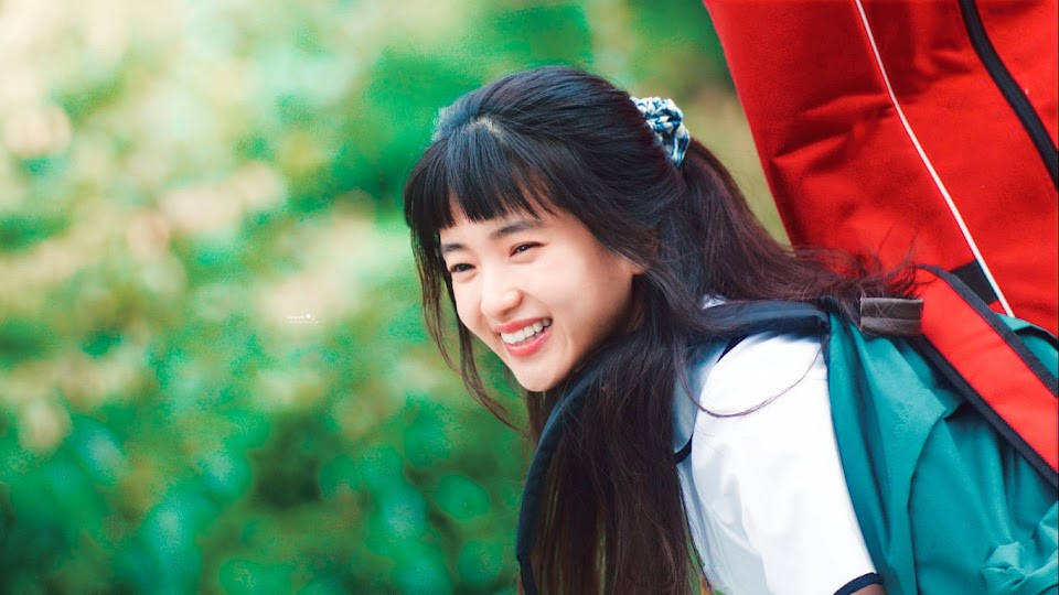 Teenage Girl 桌布 壁紙 — 一位快樂的深色頭髮年輕女性，背著紅色和藍綠色的背包，在柔和的綠色模糊背景上回頭微笑。