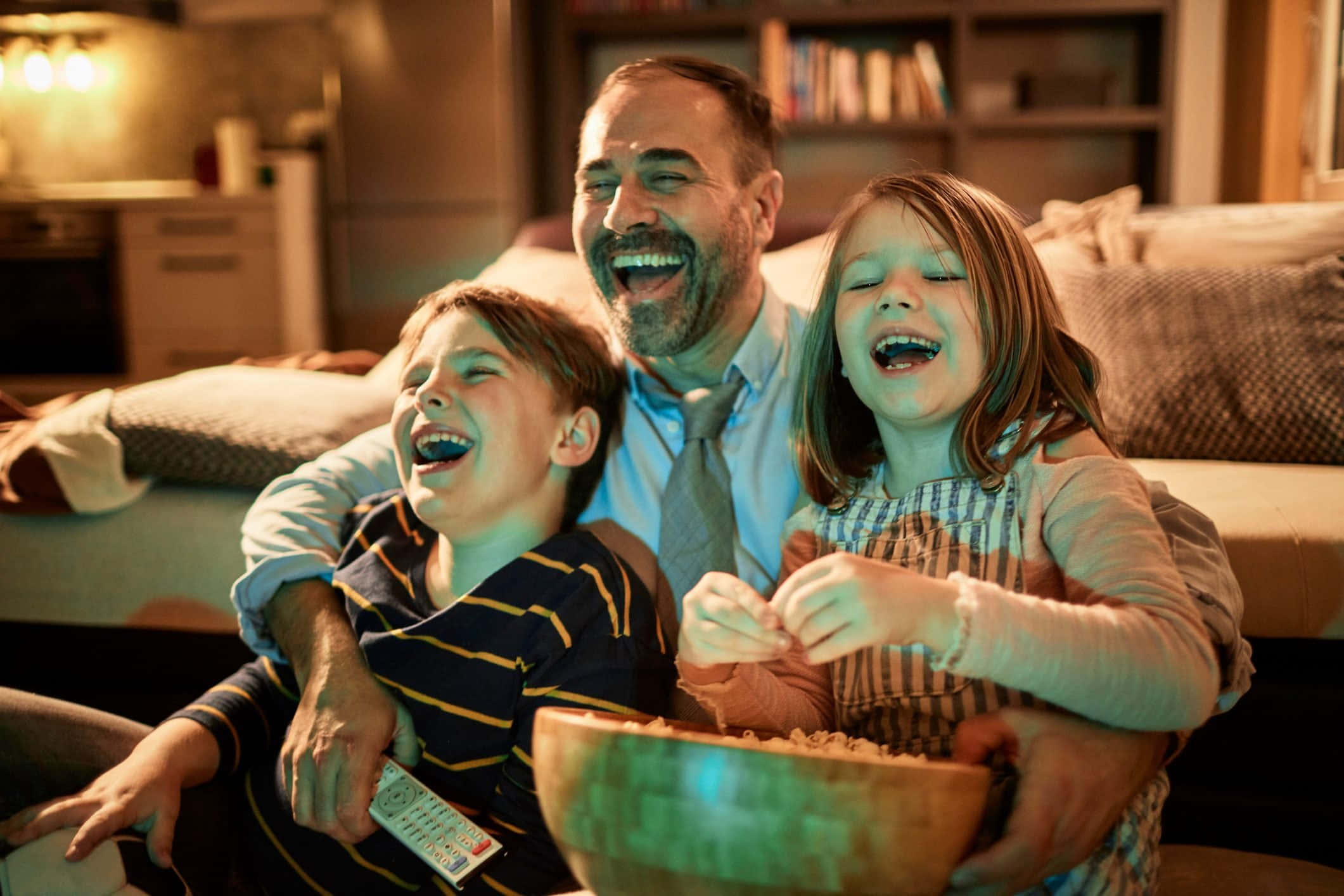 Family 桌布 壁紙 — 一位快樂的父親和他的兩個大笑的孩子在溫暖照明的客廳沙發上分享爆米花。