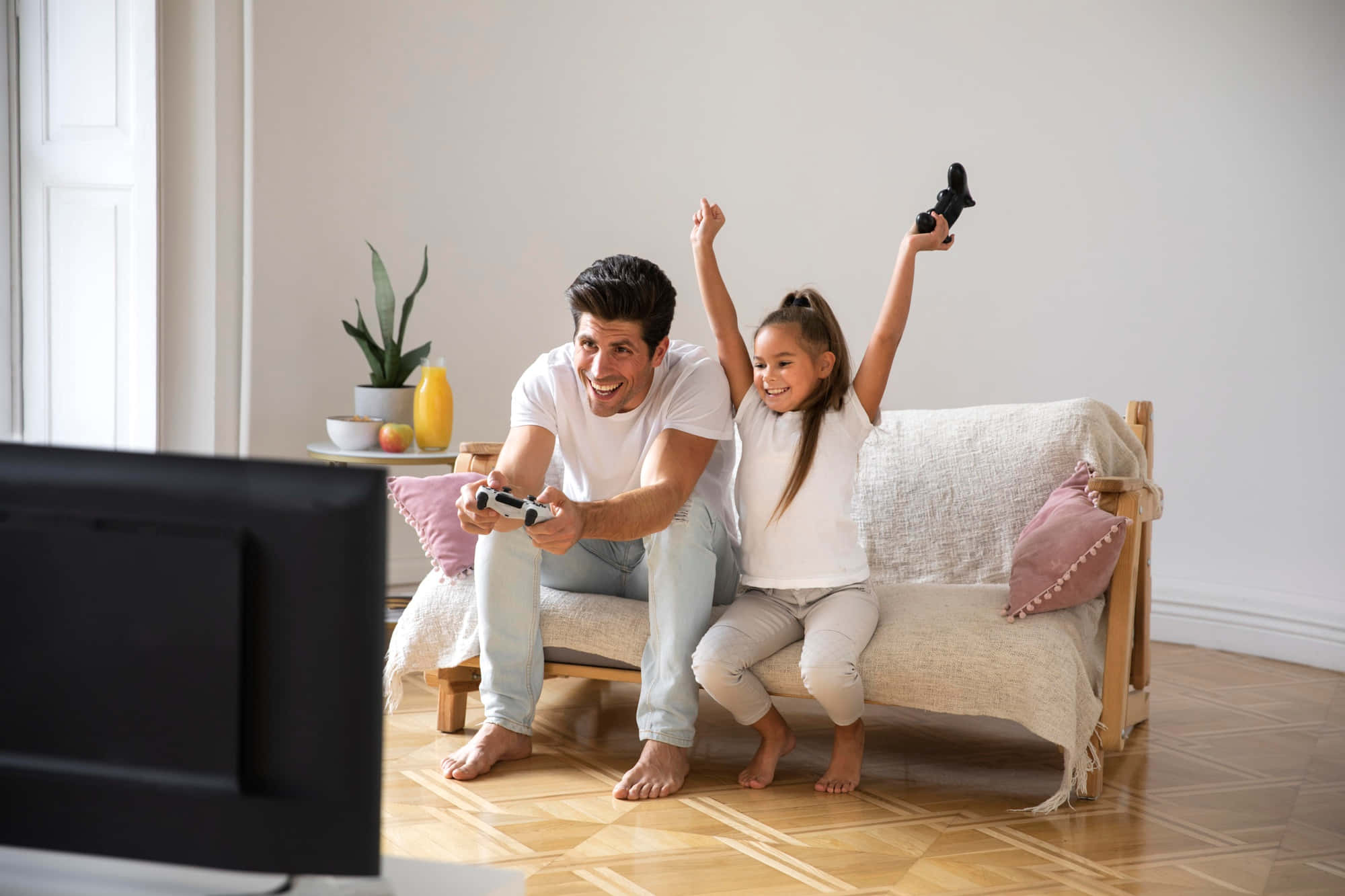 Home Entertainment 桌布 壁紙 — 一位快樂的父親和女兒在陽光充足的客廳舒適沙發上慶祝遊戲勝利。