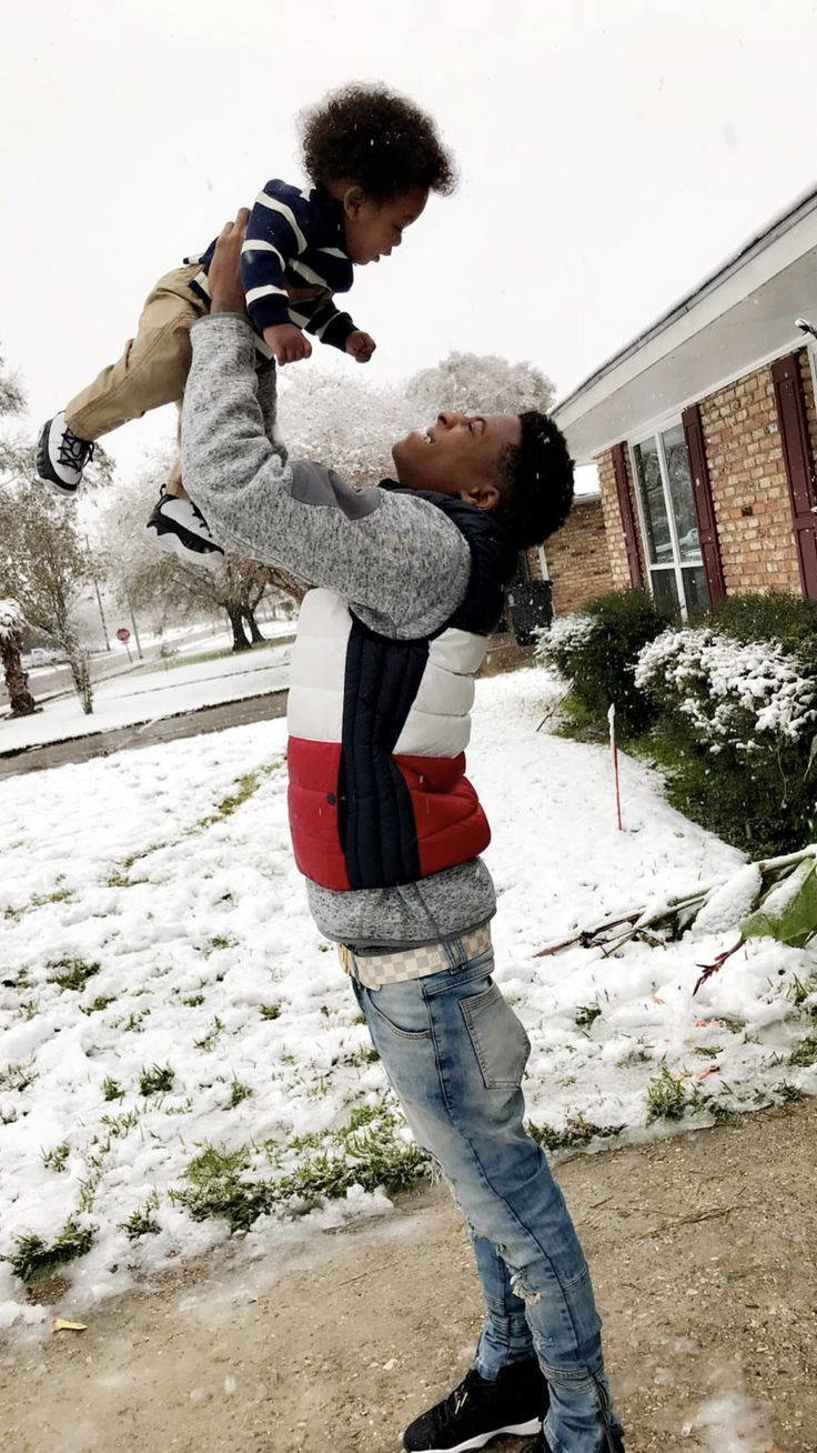 Youngboy 桌布 壁紙 — 一位快樂的父親在雪地庭院中高舉他的幼兒，雪花在他們周圍飄落。