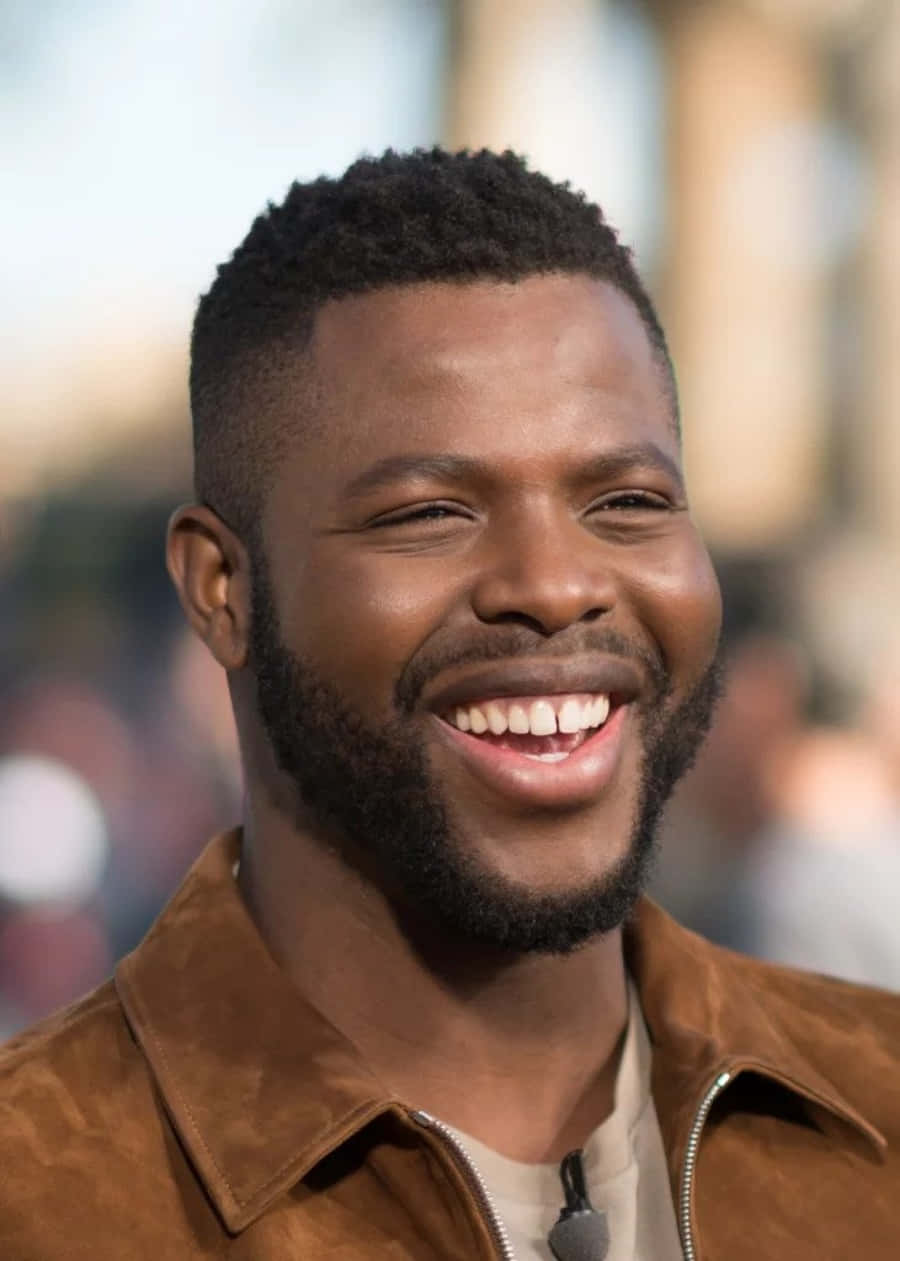 Winston Duke 桌布 壁紙 — 一位快樂的男子，帶著溫暖的笑容和短鬍鬚，穿著棕色麂皮外套，背景是柔和模糊的城市景觀。