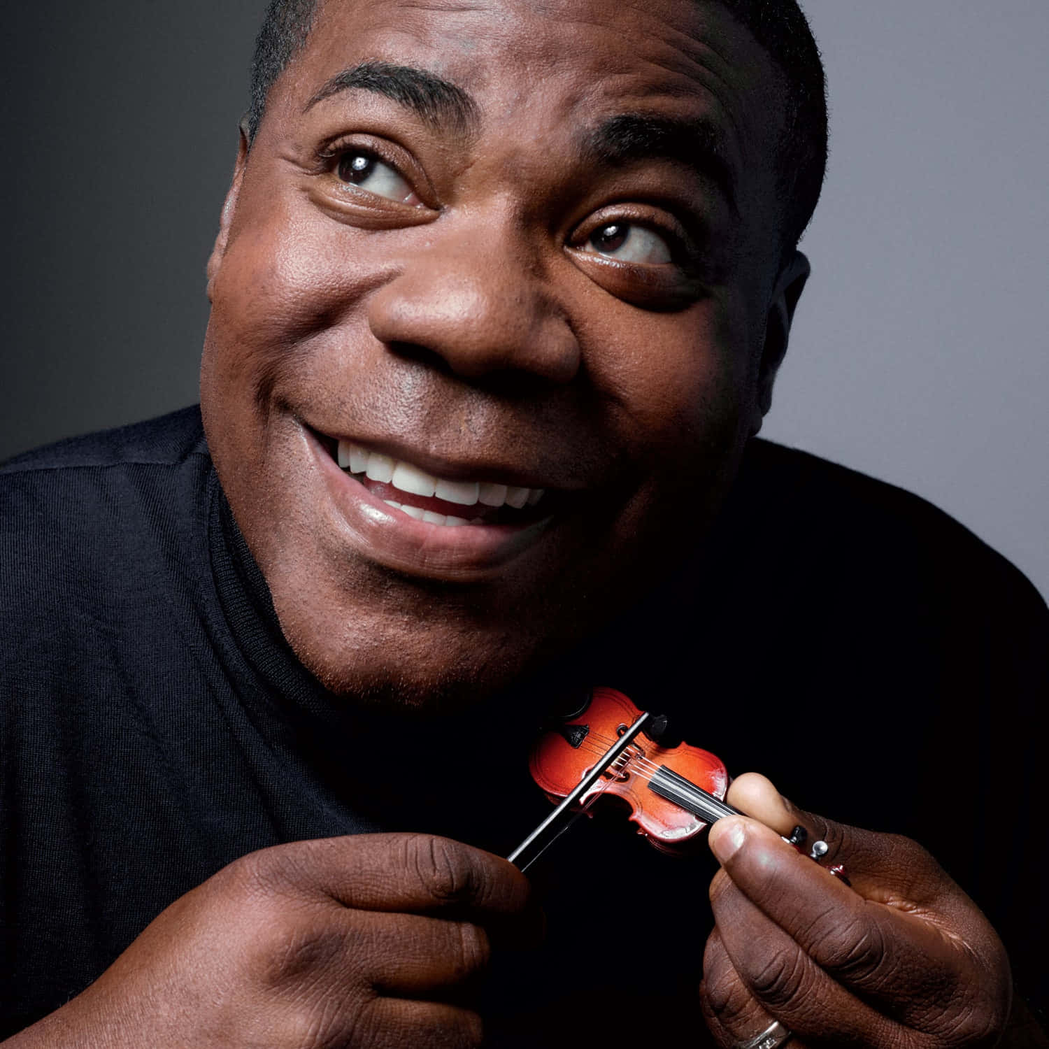 Tracy Morgan 桌布 壁紙 — 一位快樂的男子帶著溫暖的笑容，在柔和的灰色背景前拿著一個微型小提琴。