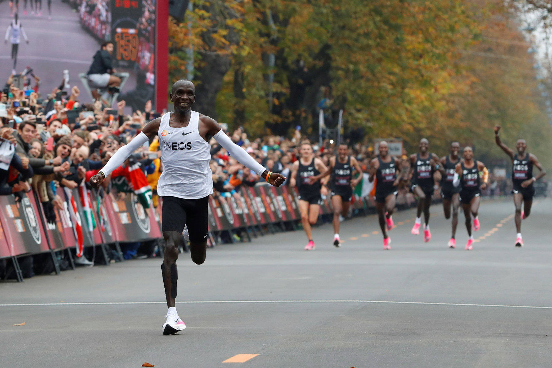 Marathon 桌布 壁紙 — 一位快樂的男性跑者張開雙臂穿過街道終點線，觀眾歡呼，秋樹排列在路線兩旁。