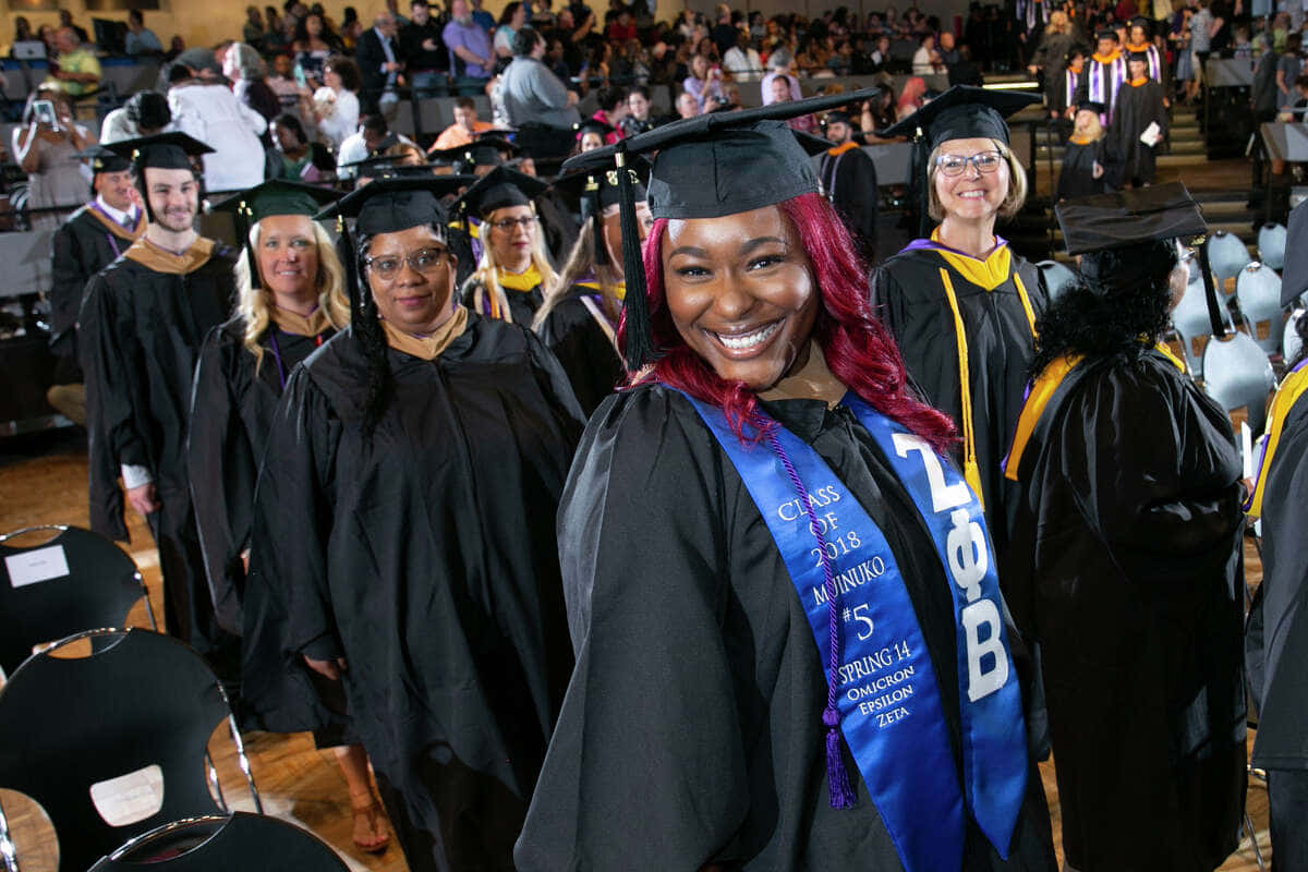 Graduation 桌布 壁紙 — 一位快樂的畢業生，擁有鮮豔的紅髮，在擁擠的禮堂中，同學們走在她身後，她戴著學士帽和長袍，容光煥發。