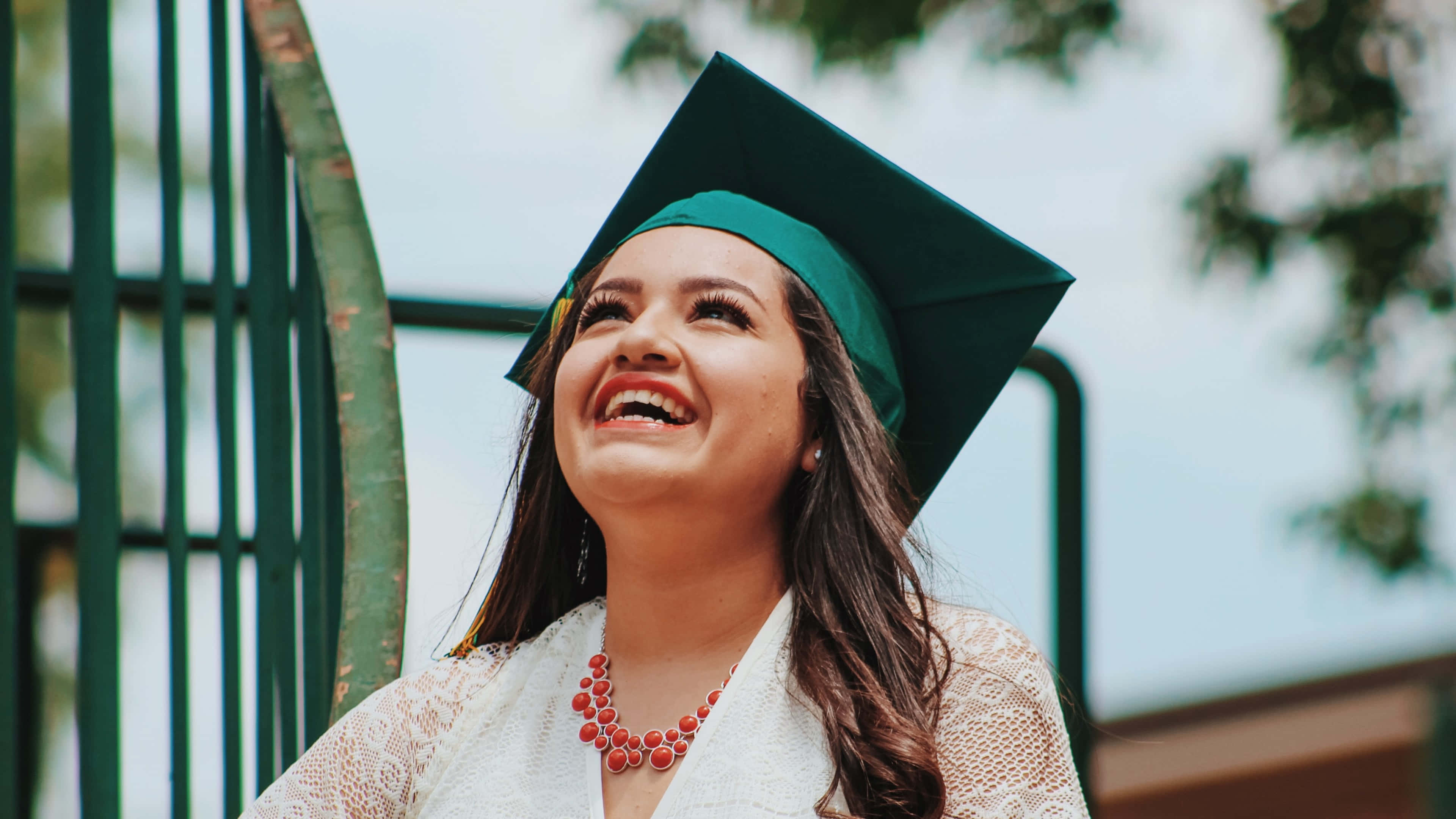 Graduation Ceremony 桌布 壁紙 — 一位快樂的畢業生戴著綠色四方帽和白色畢業袍，在戶外樹木和圍欄背景下開懷大笑。