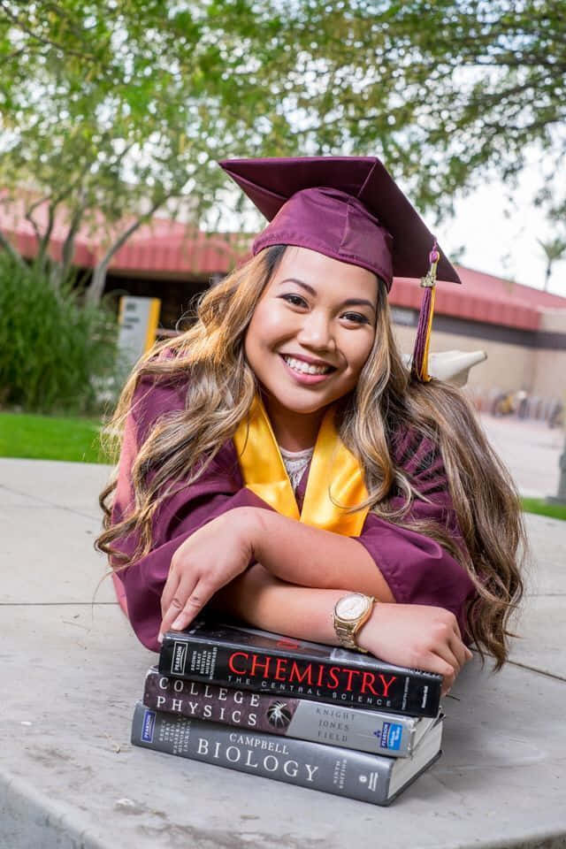 Graduation 桌布 壁紙 — 一位快樂的畢業生身穿栗色學士袍和四方帽，微笑著靠在標有化學、物理和生物學的教科書上。