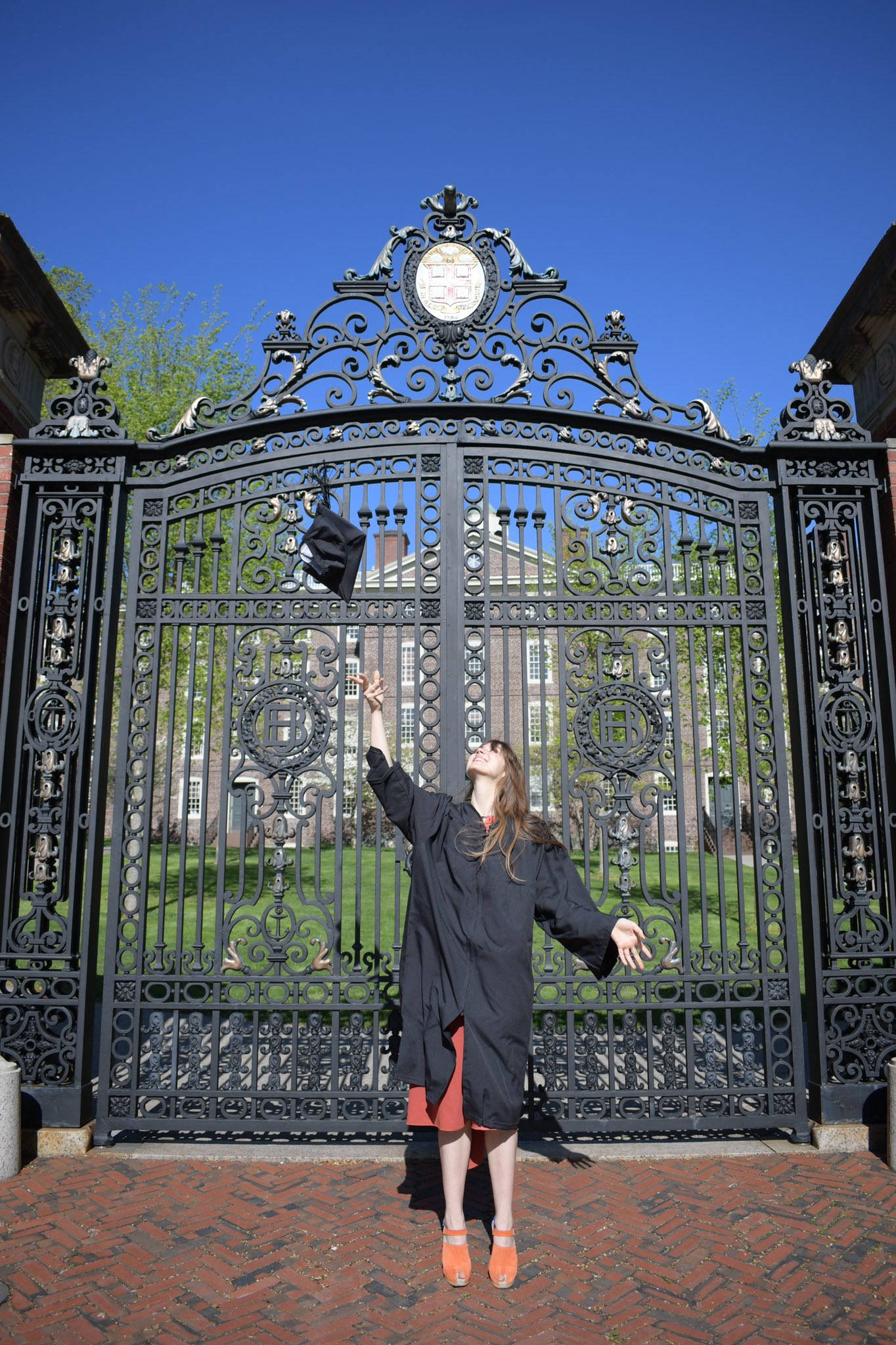 Graduation 桌布 壁紙 — 一位快樂的畢業生身穿黑色長袍和橙色鞋子，在晴朗的藍天下將帽子高高拋過華麗的鍛鐵大門。