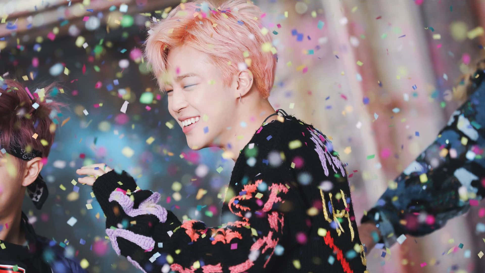 K-pop 桌布 壁紙 — 一位快樂的粉紅色頭髮年輕男子在色彩繽紛的紙屑如雨般落在他周圍的熱鬧慶祝活動中大笑。