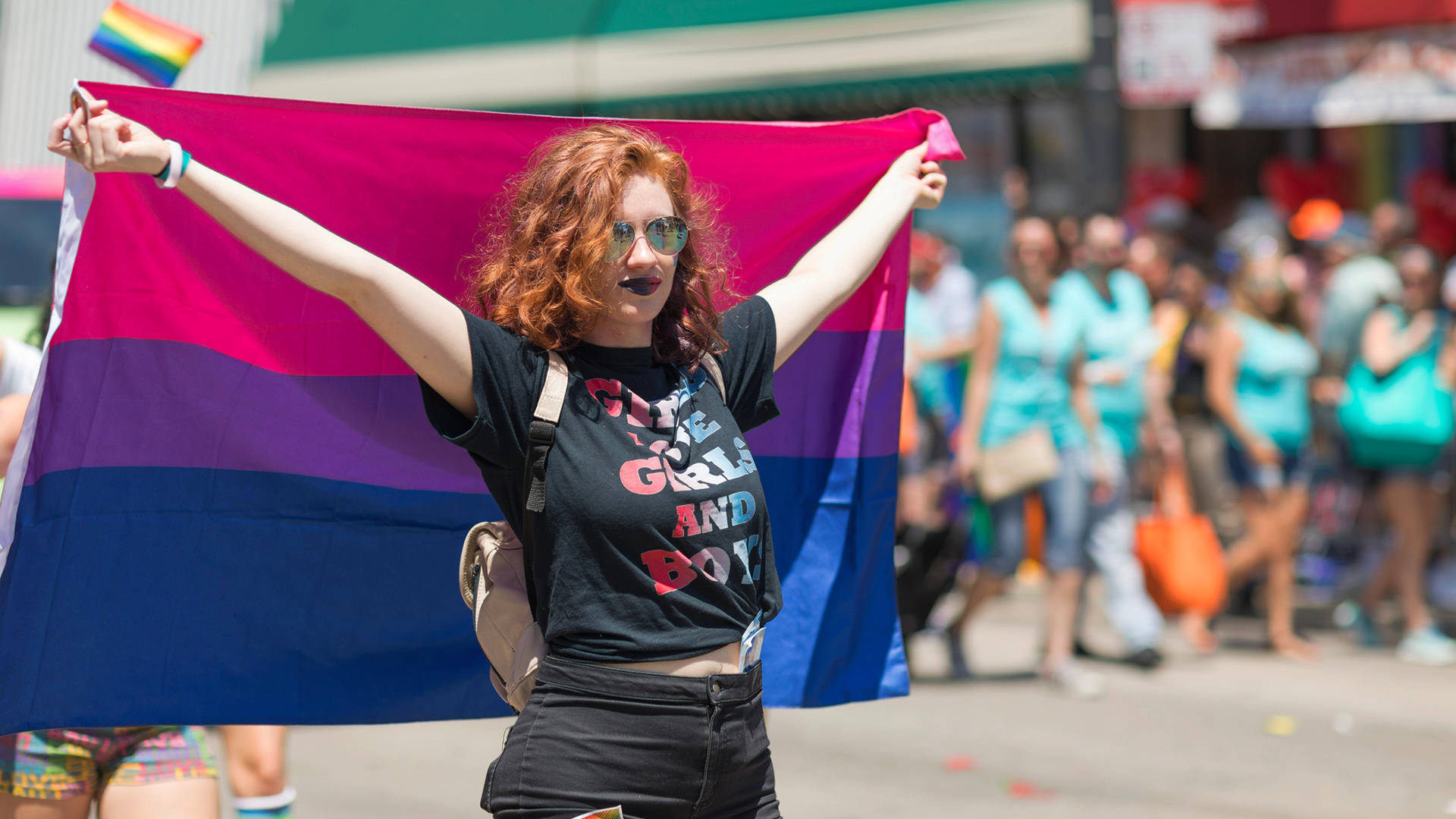 Bisexual Flag 桌布 壁紙 — 一位快樂的紅髮女子戴著太陽眼鏡，在熱鬧的街頭遊行中自豪地高舉雙性戀驕傲旗幟。