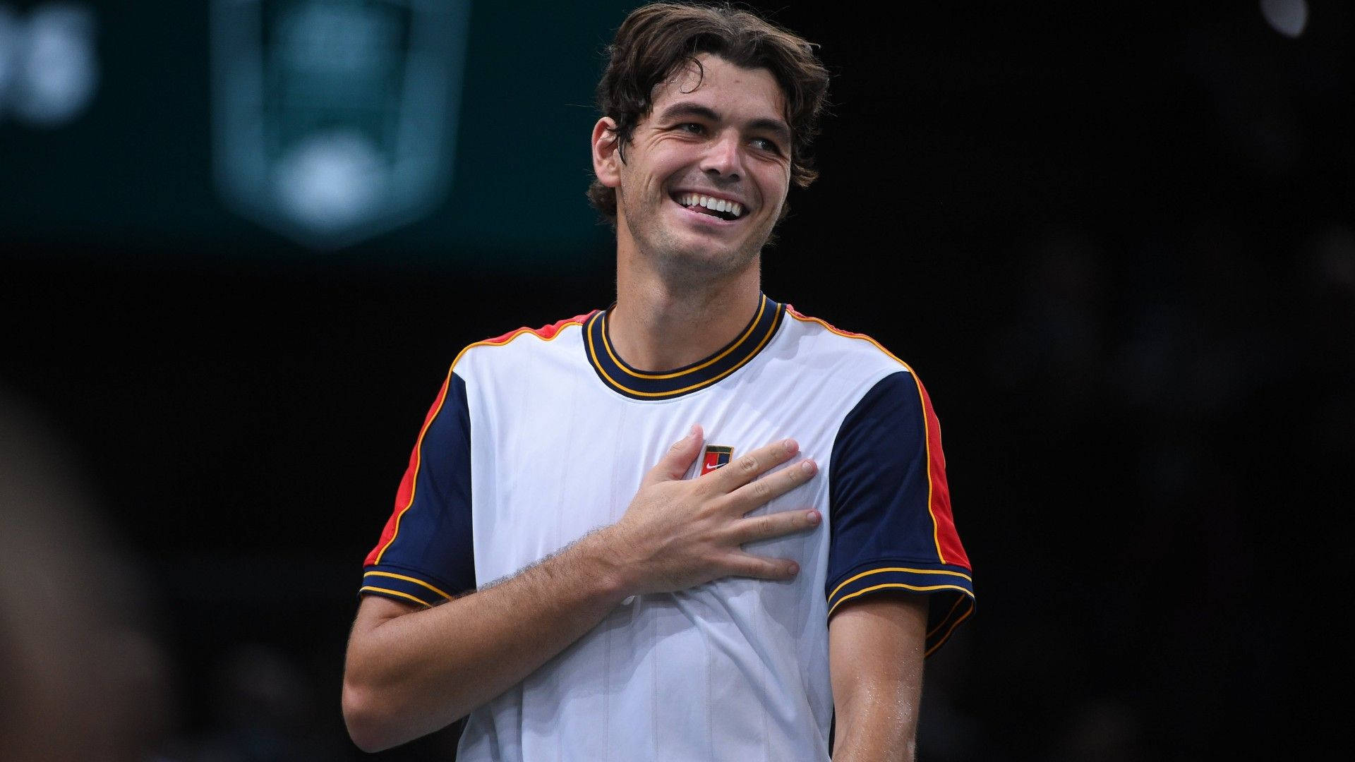 Tennis 桌布 壁紙 — 一位快樂的網球選手在昏暗的場館中，在明亮的聚光燈下，手放在心口，洋溢著自豪的笑容。