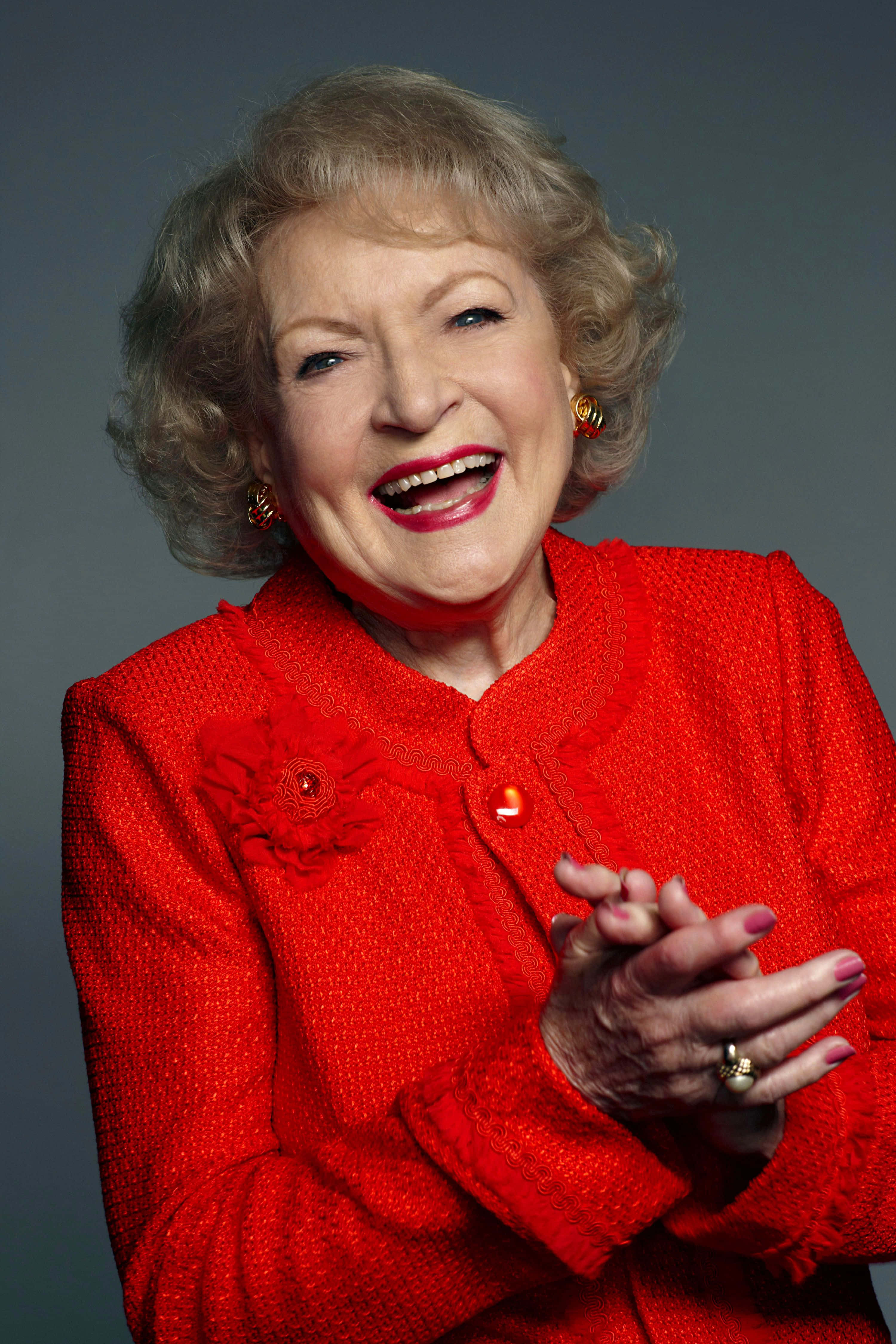 Betty White 桌布 壁紙 — 一位快樂的老年女士穿著鮮紅色紋理外套，在純灰色攝影棚背景前雙手合十開懷大笑。