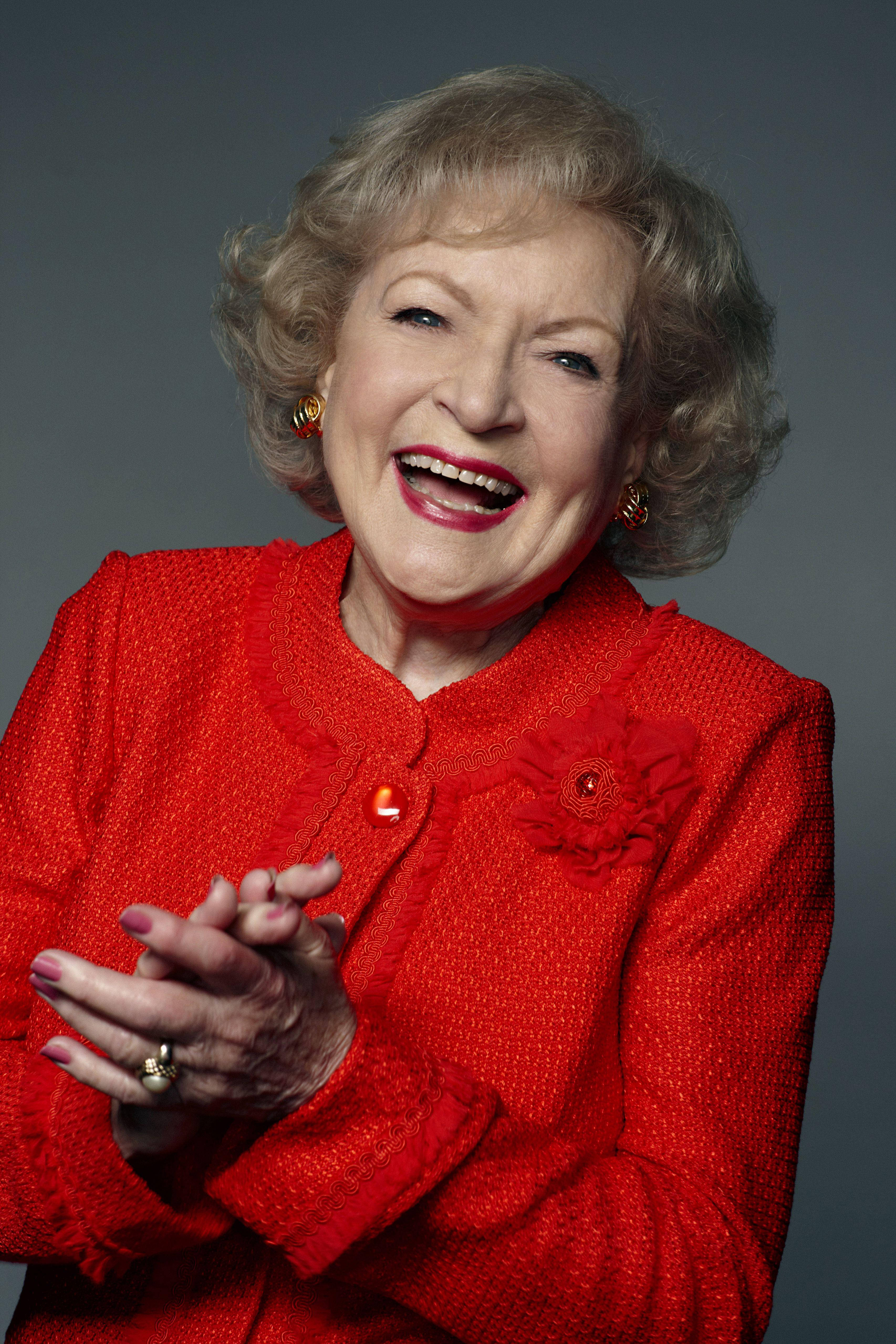 Betty White 桌布 壁紙 — 一位快樂的老年女士穿著鮮紅色質感外套，在純灰色工作室背景前雙手合十開懷大笑。